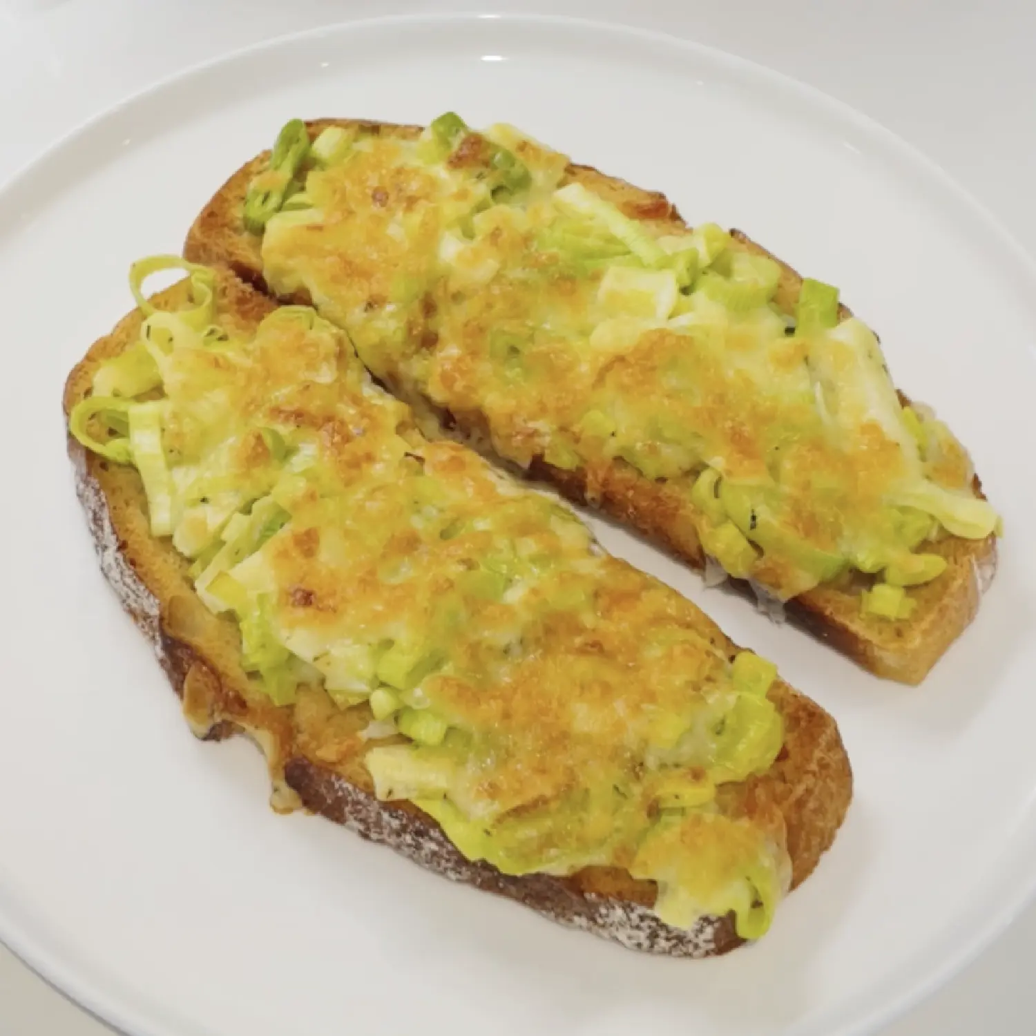 Two slices of crusty toasted bread on a white plate, topped with melted golden cheese and sliced green leeks.