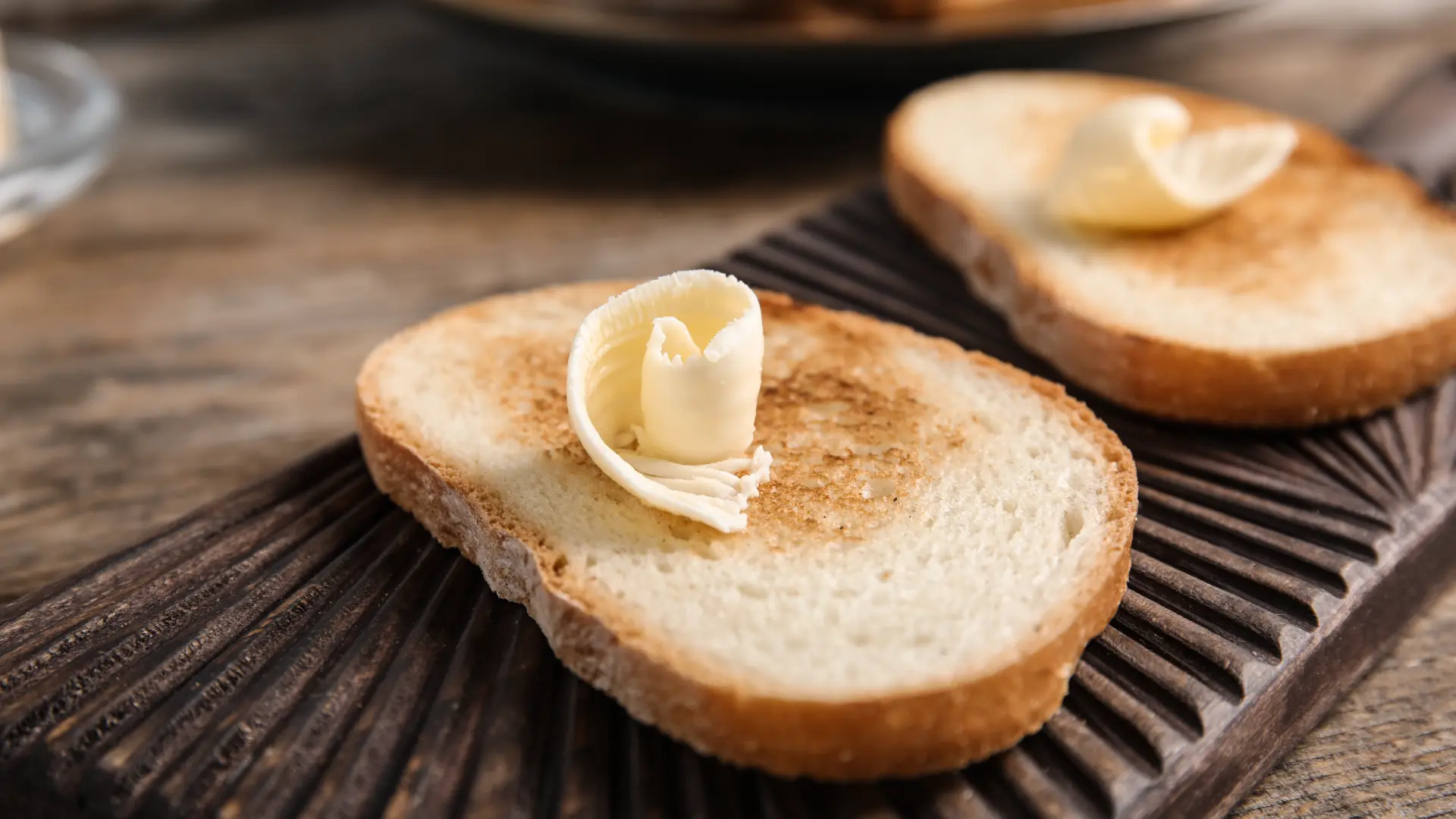 Two slices of toasted bread, each topped with a decorative curl of butter, resting on a dark, textured wooden board.