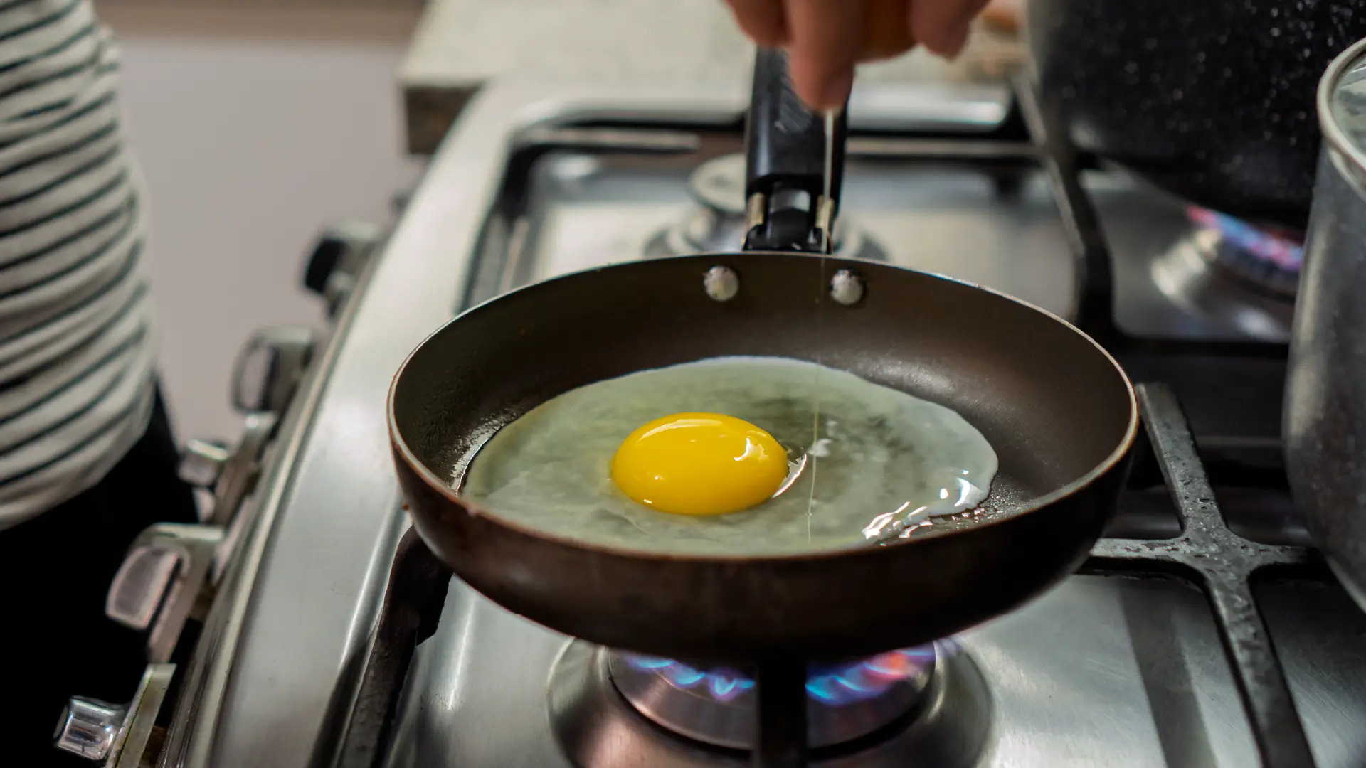 A raw egg cooking in a small frying pan over a blue gas flame stove, with a hand seen just above the pan.