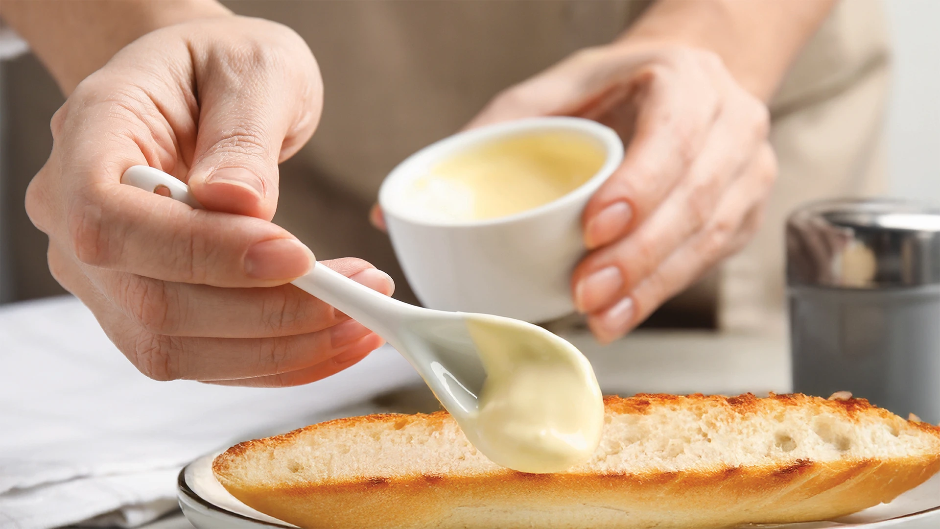 Someone spreads mayonnaise with a spoon onto the bottom half of a toasted sub roll
