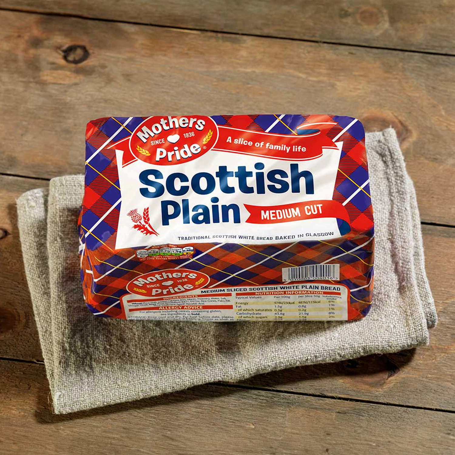 Top-down view of a pack of Mothers Pride Scottish Plain bread in tartan packaging, placed on a beige linen cloth covering a rustic wooden table.