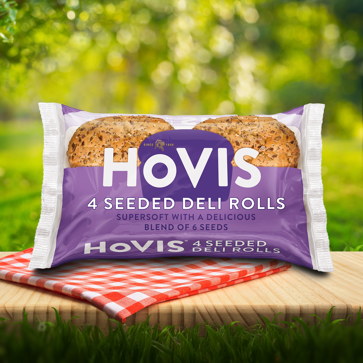 A pack of Hovis 4 Seeded Deli Rolls packaging on a picnic table with a green outdoor background. The pack shows seeded deli rolls  and highlights “Supersoft with a delicious blend of 6 seeds.” A red gingham cloth adds a picnic feel to the setting.