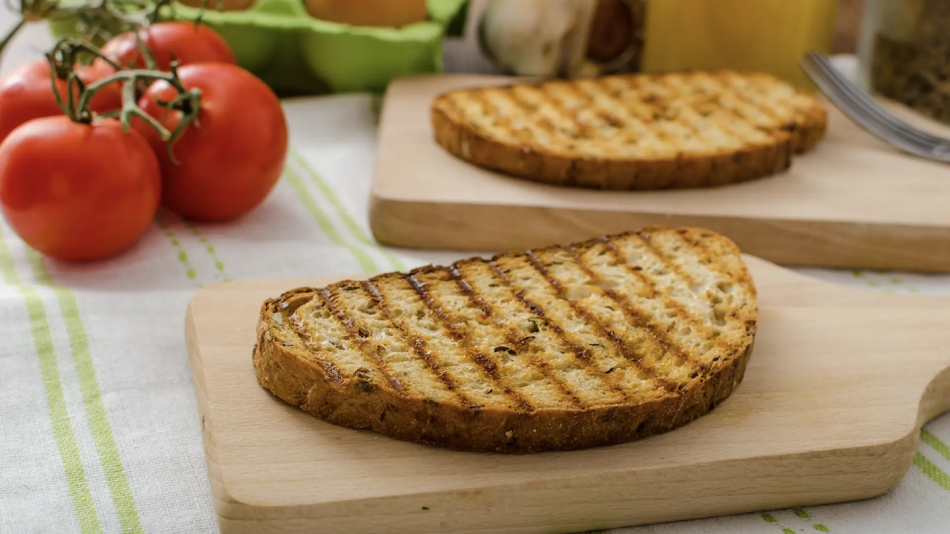 Two grilled bread slices with char marks on small wooden boards, with fresh red vine tomatoes in the background.