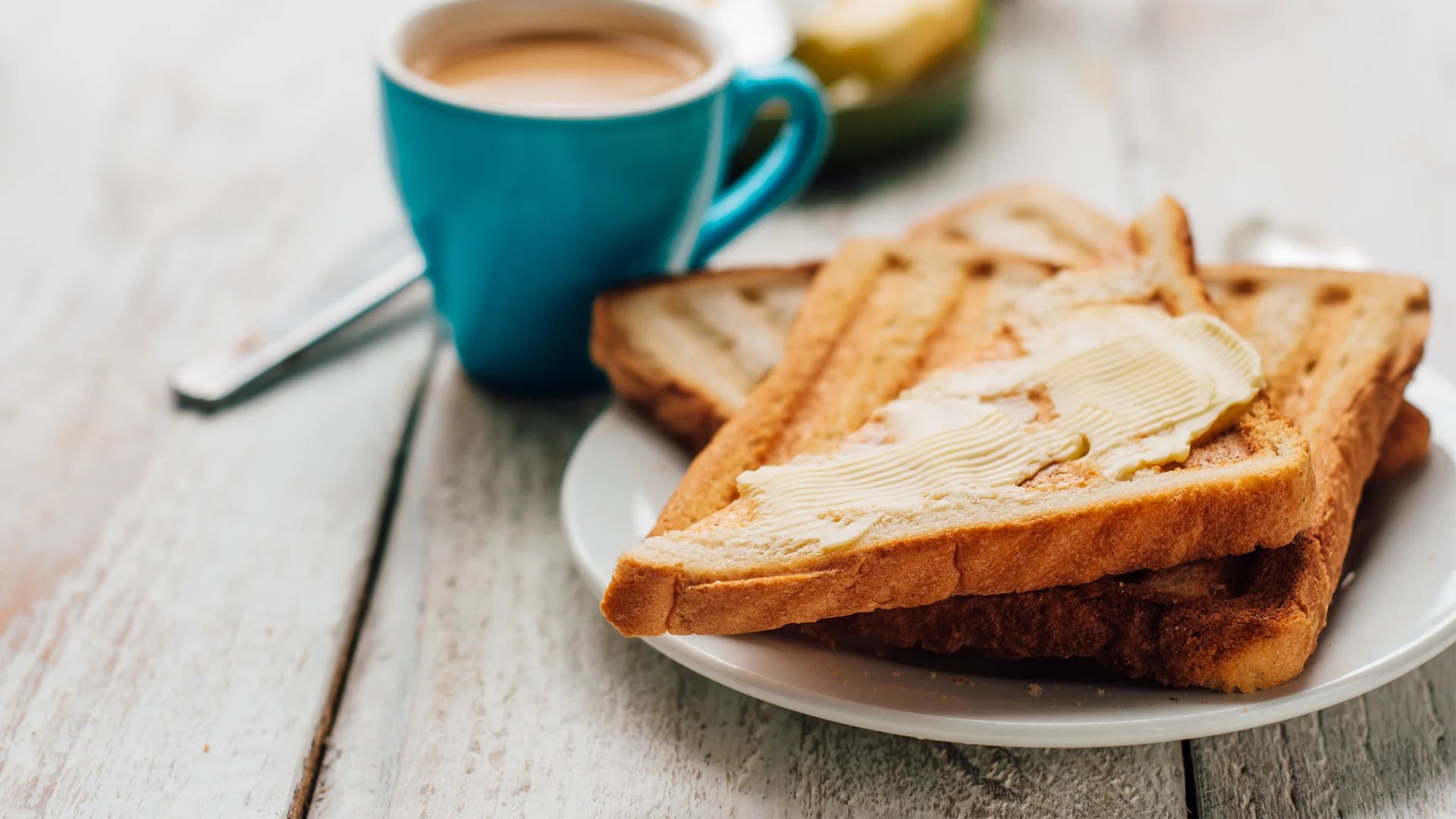 A pile of buttered toast sits on a wooden table with a cup of tea nearby. 