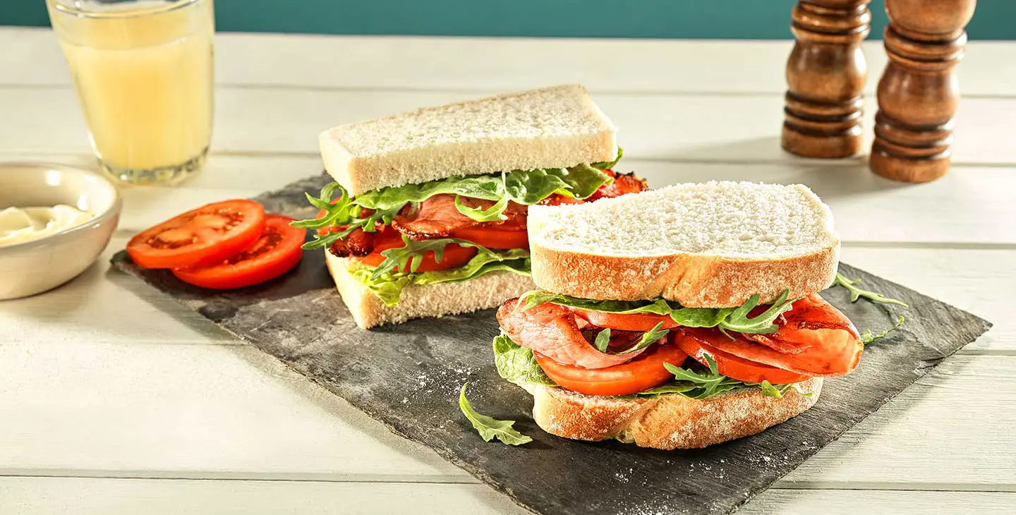 A bacon, lettuce and tomato sandwich is placed on a piece of slate on a kitchen tables. 