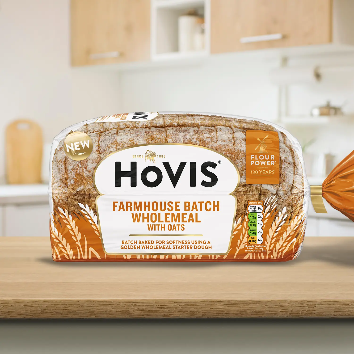 A loaf of Farmhouse Batch Wholemeal with Oats bread with sourdough and a crust dusted with flour, displayed on a wooden kitchen countertop. The packaging features a white and orange design with a gold trim and nutrition information. A 'NEW' label is shown on the top corner of the pack