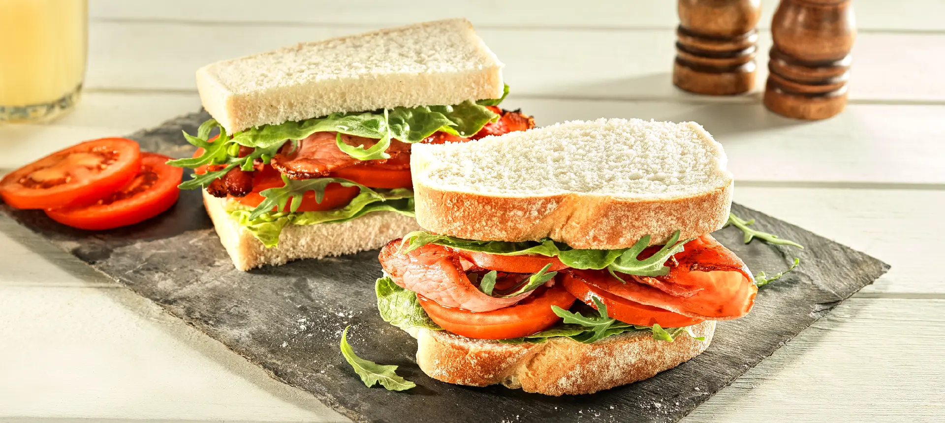 A sandwich made with Hovis Farmhouse Soft White cut in half, with crispy bacon, salad leaves and tomato slices between white sliced bread.