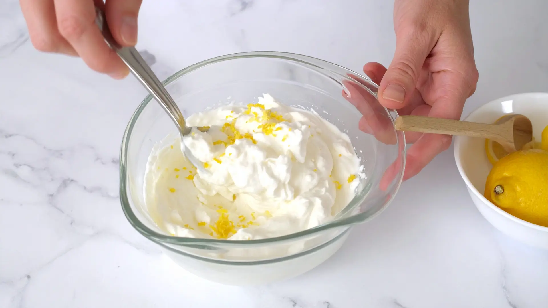 A person stirs a creamy mixture of yogurt, cream cheese, and lemon zest in a glass bowl, with lemons nearby
