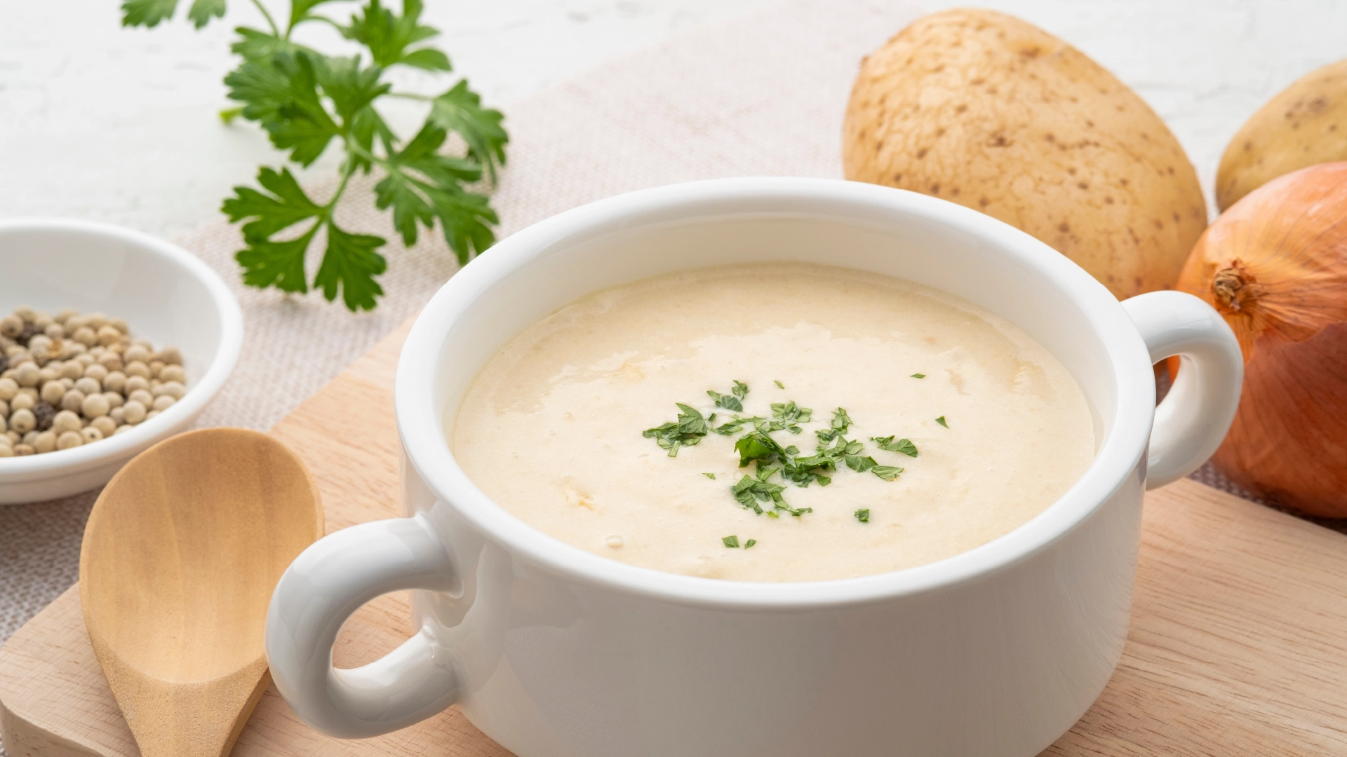 A white bowl filled with creamy beige potato soup, topped with sliced parsley, sits on a wooden chopping board. In the background are two potatoes, an onion, and some parsley leaves.