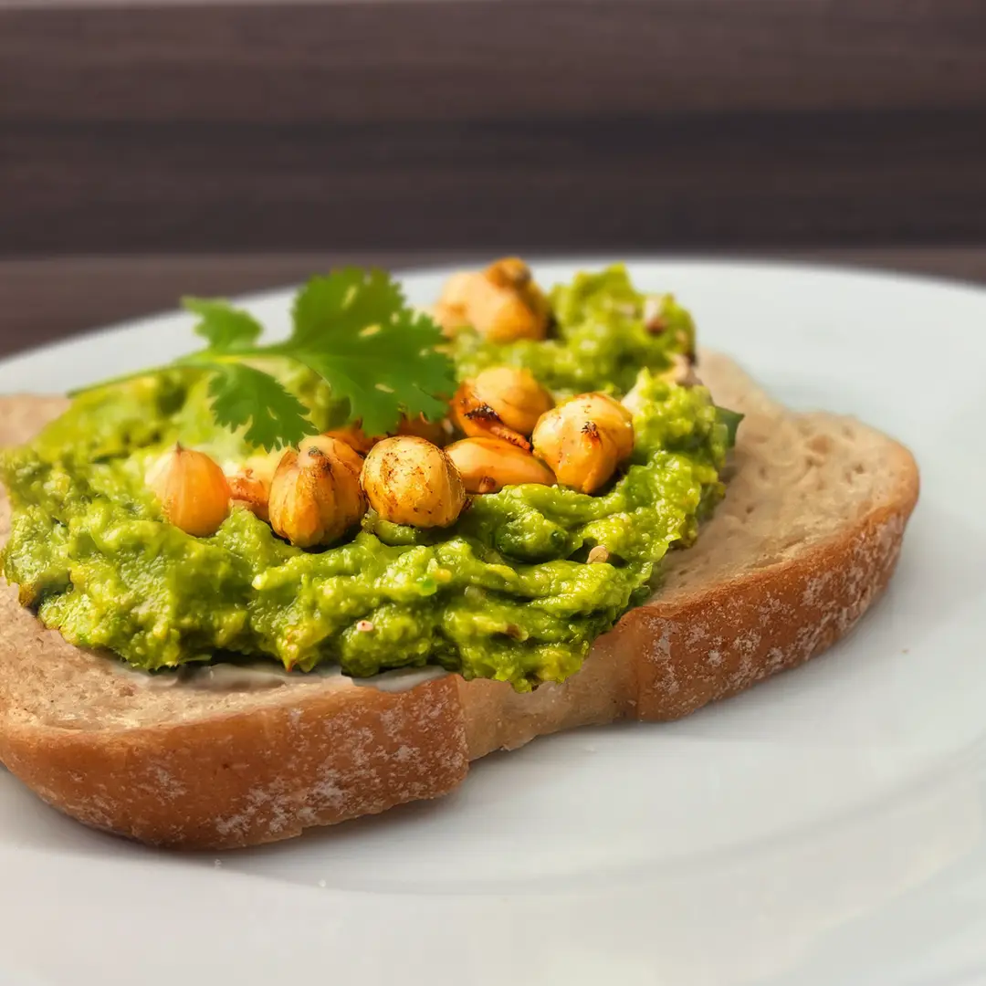 A close-up, eye-level shot of a slice of wholemeal toast on a white plate, generously topped with vibrant green smashed avocado and chickpeas. The chickpeas are lightly browned and seasoned, adding texture. A fresh sprig of cilantro garnishes the topping. The background is a blurred dark wood surface.