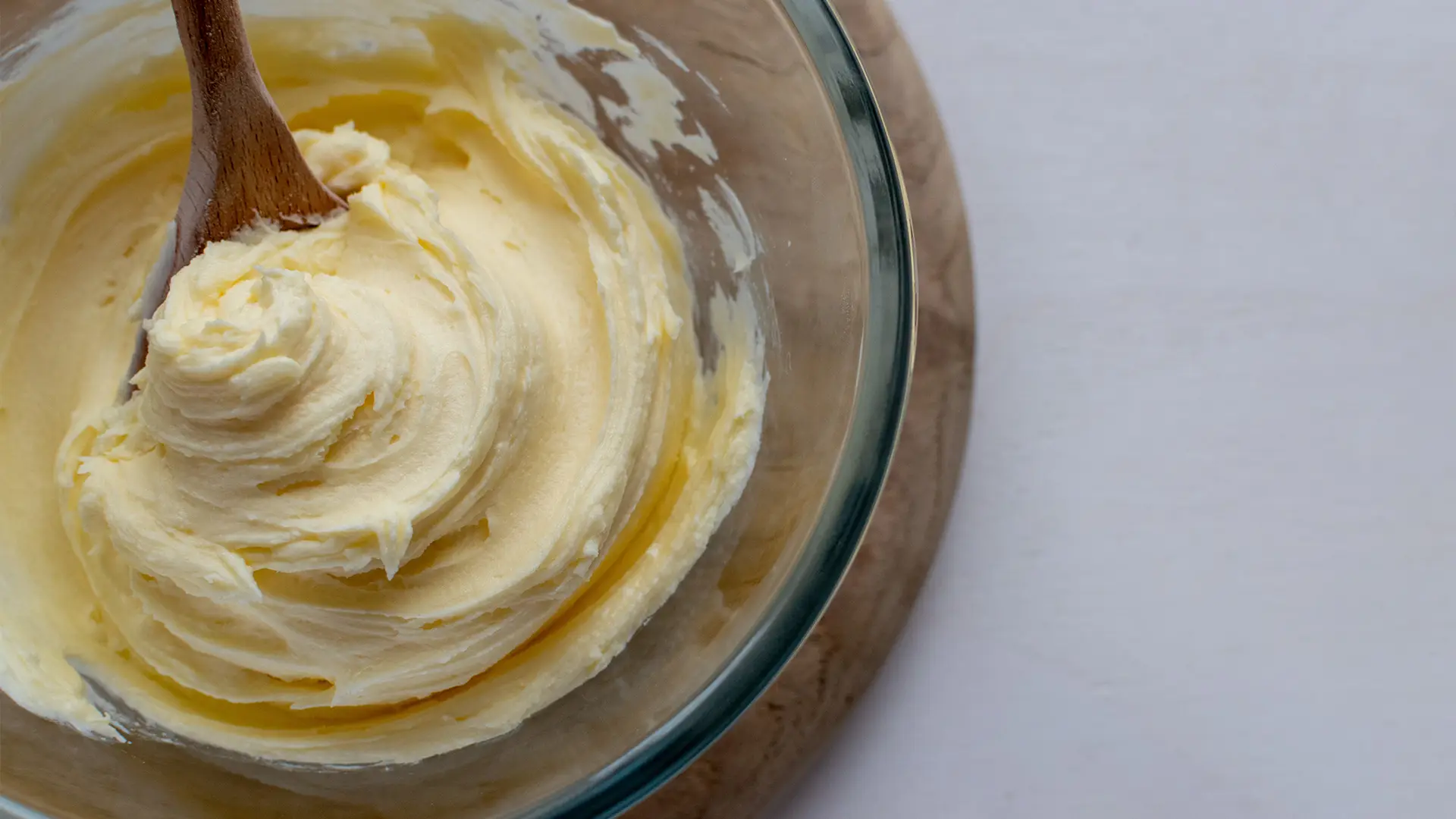 A close-up, top-down view shows a clear glass bowl filled with creamy, pale yellow frosting or butter mixture, deeply swirled by a wooden spoon. The spoon rests in the bowl's centre, creating a prominent swirl pattern in the mixture. The bowl is set on a light brown wooden cutting board, which is partially visible on the left side of the frame. The rest of the background is a plain white surface.