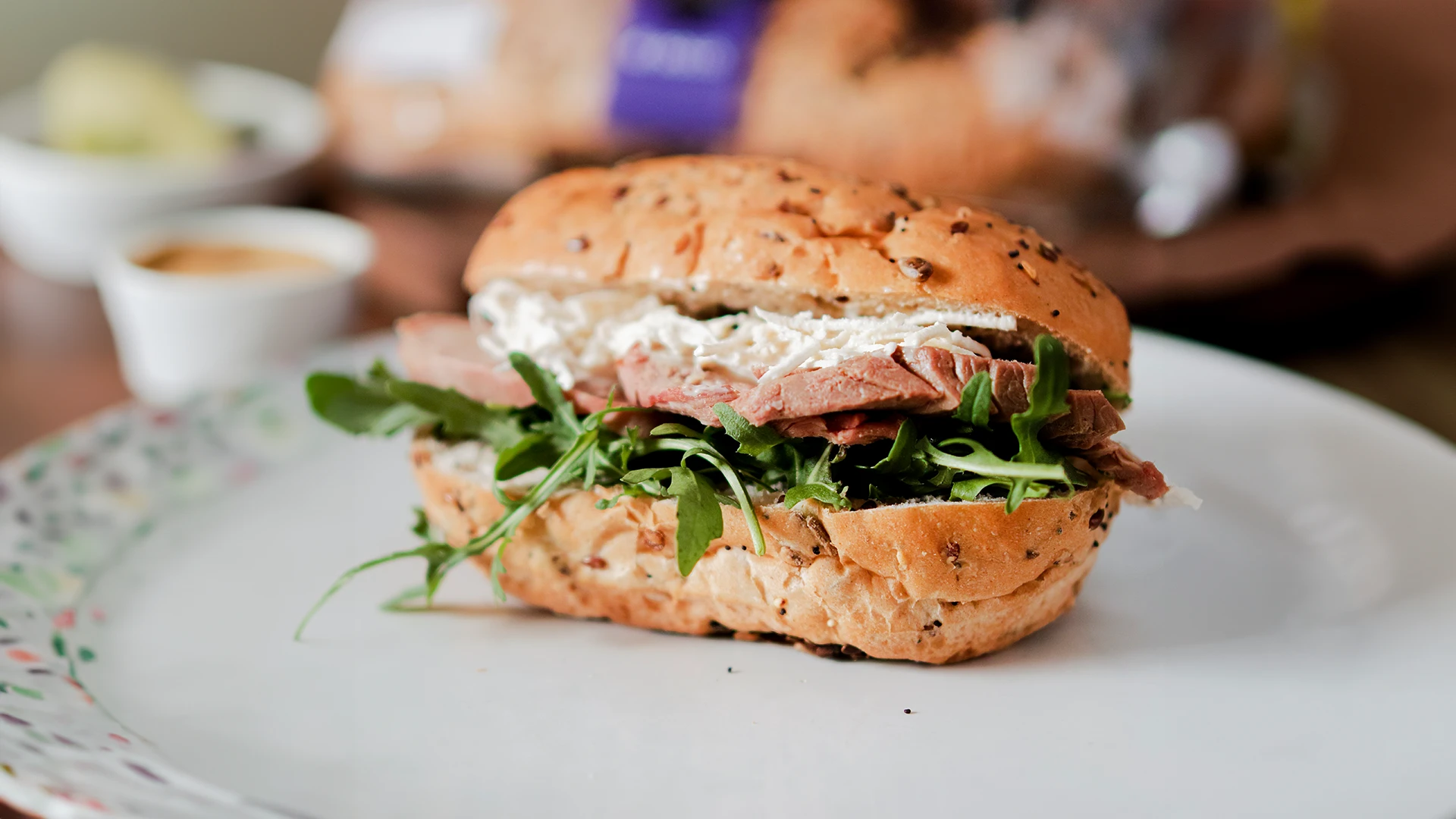 A sandwich made with Hovis 4 Seeded Deli Rolls, filled with grated cheese, Reuben beef, and fresh rocket leaves, served on a plate alongside a small bowl filled with savoury sauce.