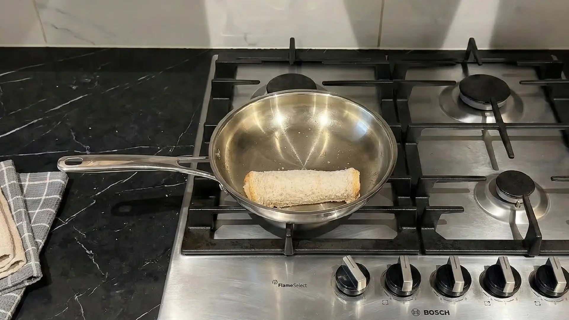 A bread roll frying in a stainless steel pan on a gas stovetop, set against a black marble countertop.
