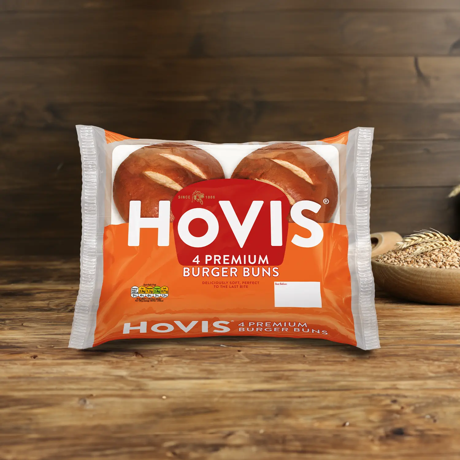 Four Hovis Burger Buns on a wooden table, with a bowl of wheat grains, a wooden scoop, and dried wheat in the background.