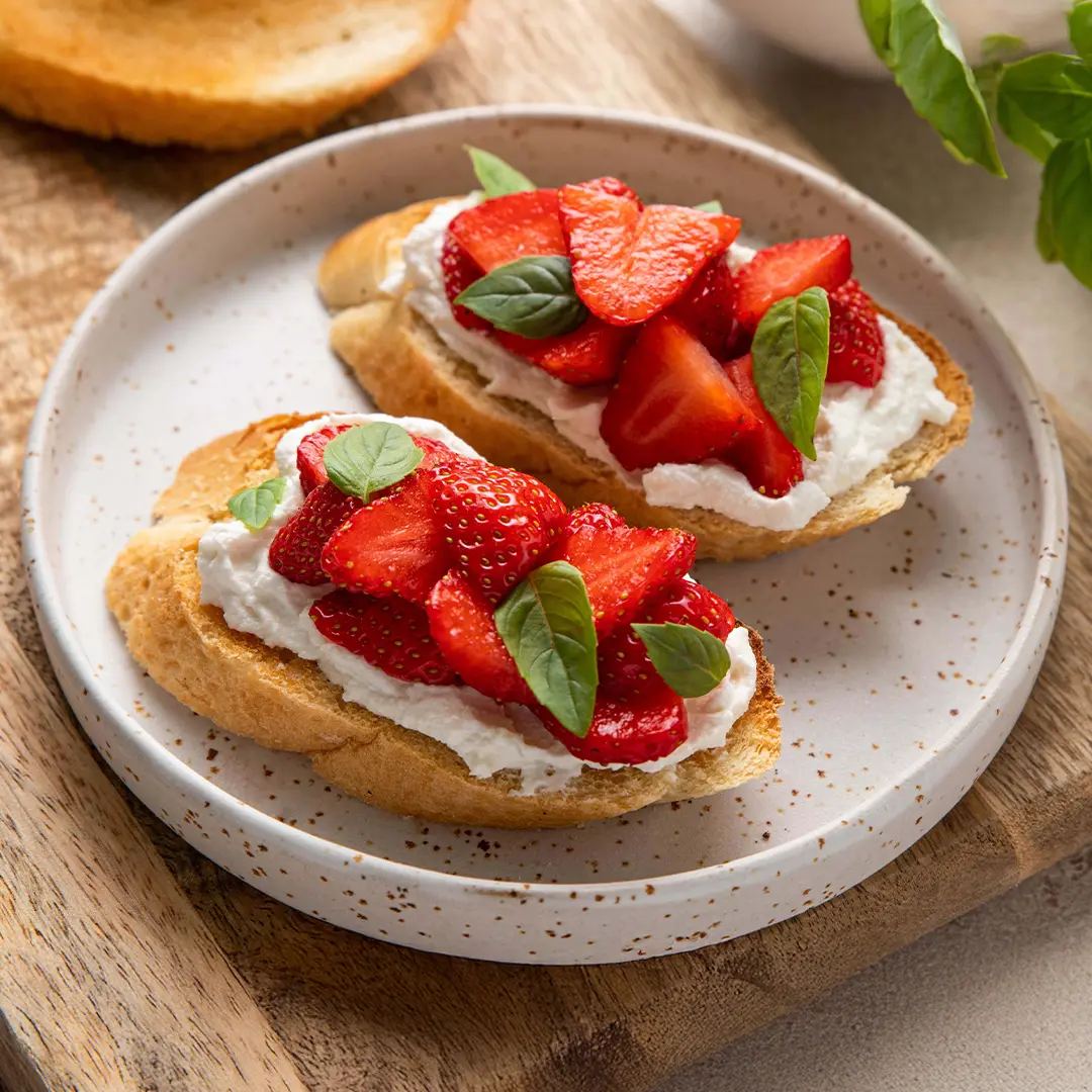 Two slices of toasted bread topped with creamy cheese, fresh strawberry slices, and basil leaves, served on a speckled ceramic plate.