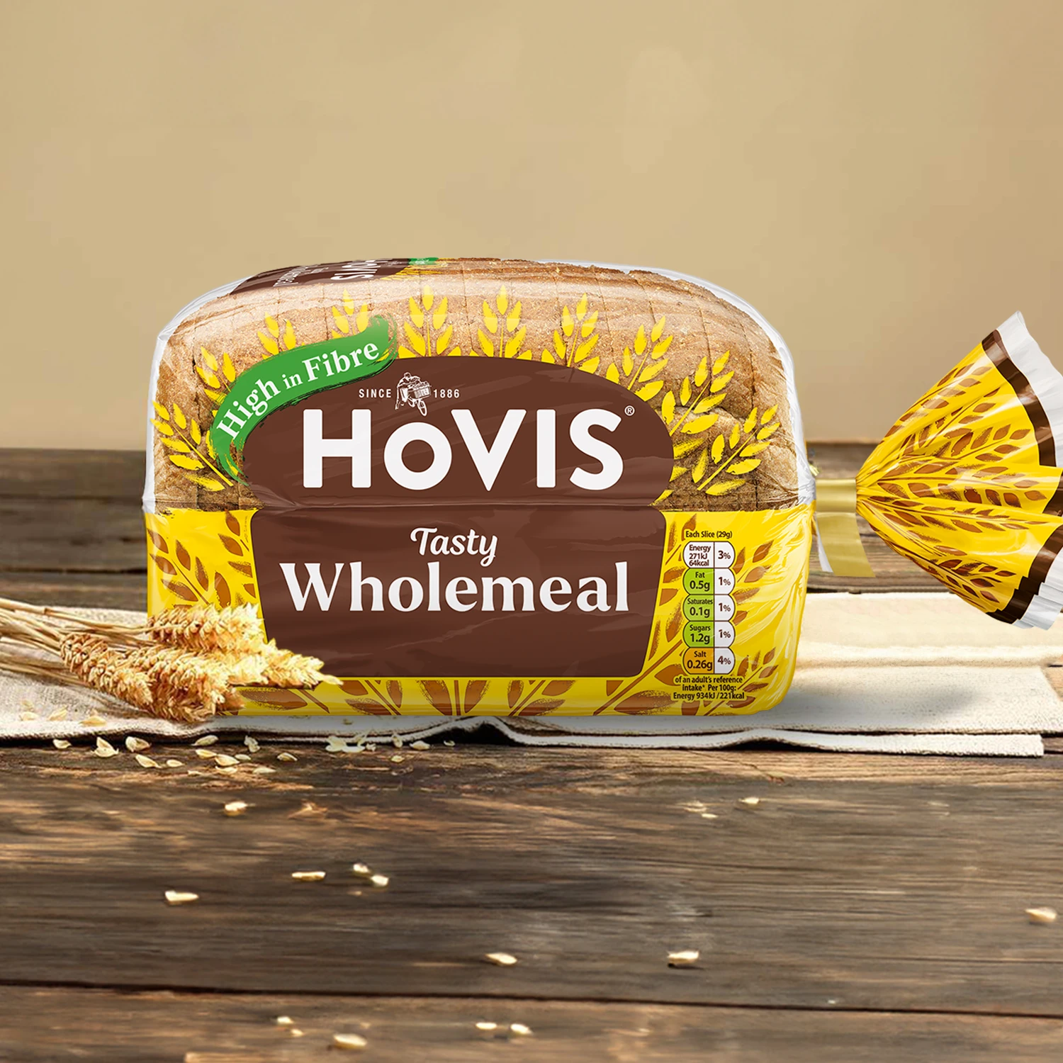 Hovis Tasty Wholemeal 400g bread on napkins against a light beige background atop a wooden table, surrounded by dried wheat.