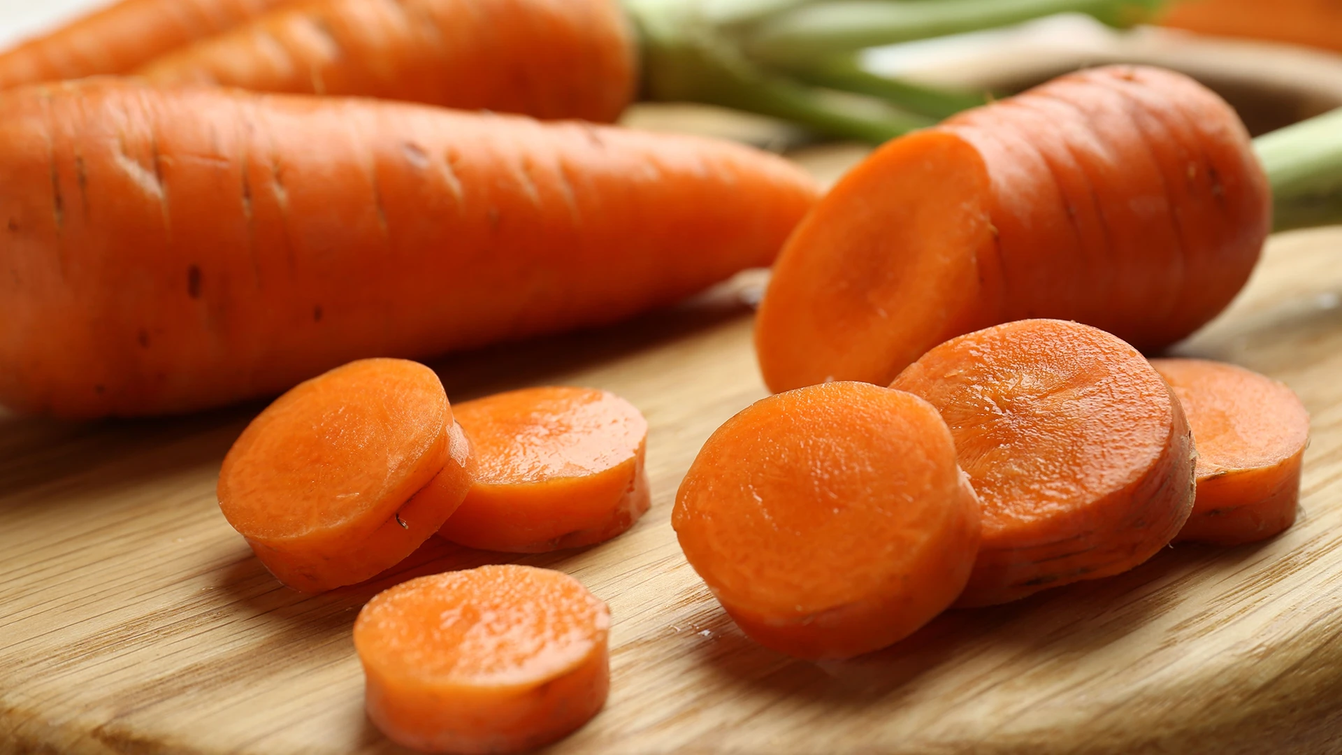 A carrot sits next to six round slices on a wooden chopping board, with extra carrots visible in the background.
