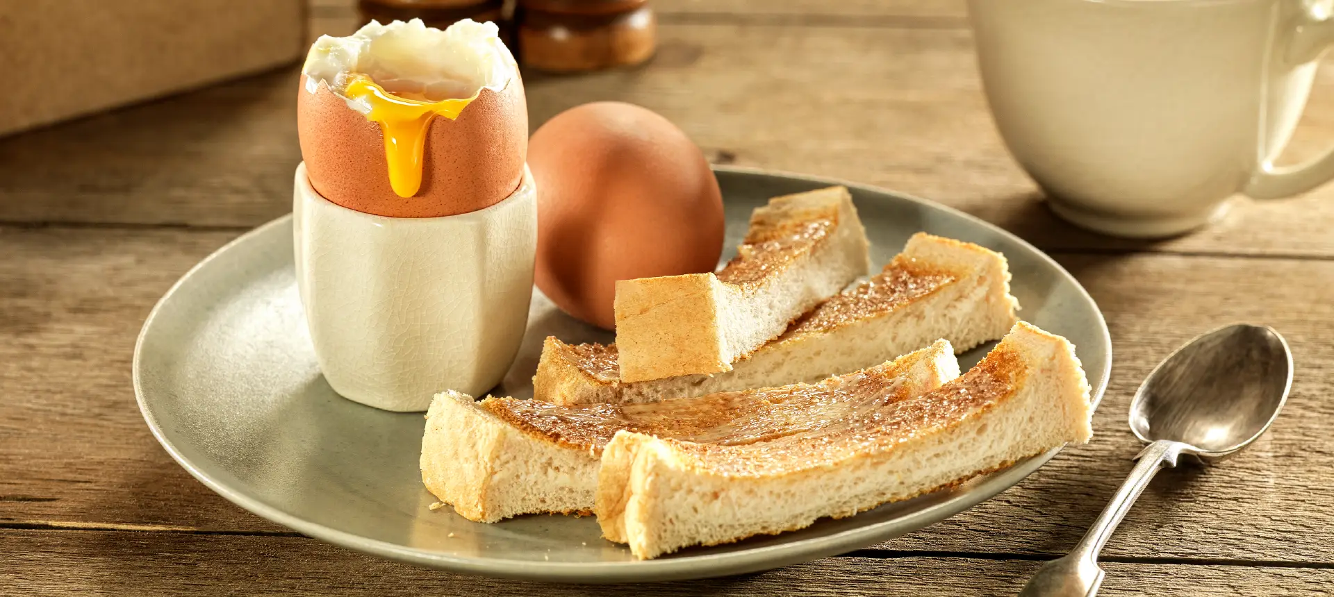 Strips of toasted Hovis Soft White Extra Thick bread alongside a soft-boiled egg with a runny yolk in an egg holder, with a spoon on the side.