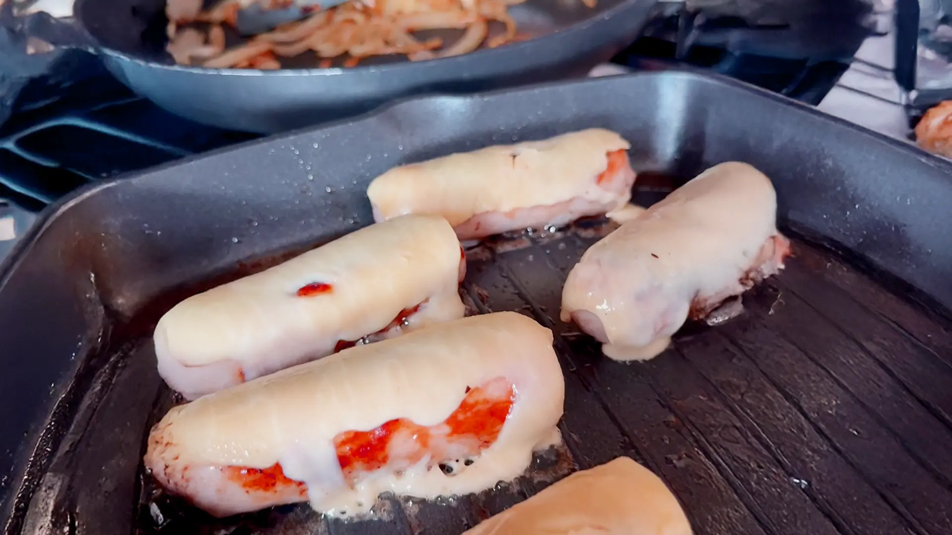 Four hot dogs cooking in a ridged black grill pan. Each hot dog is partially covered with melted, pale yellow cheese, which is bubbling and oozing. Some reddish pieces from within the hot dogs are visible where the cheese hasn't covered them. In the blurred background, another frying pan can be seen, containing what appears to be cooked onions or other ingredients. The overall scene suggests a cooking process, possibly for hot dogs or sausages.