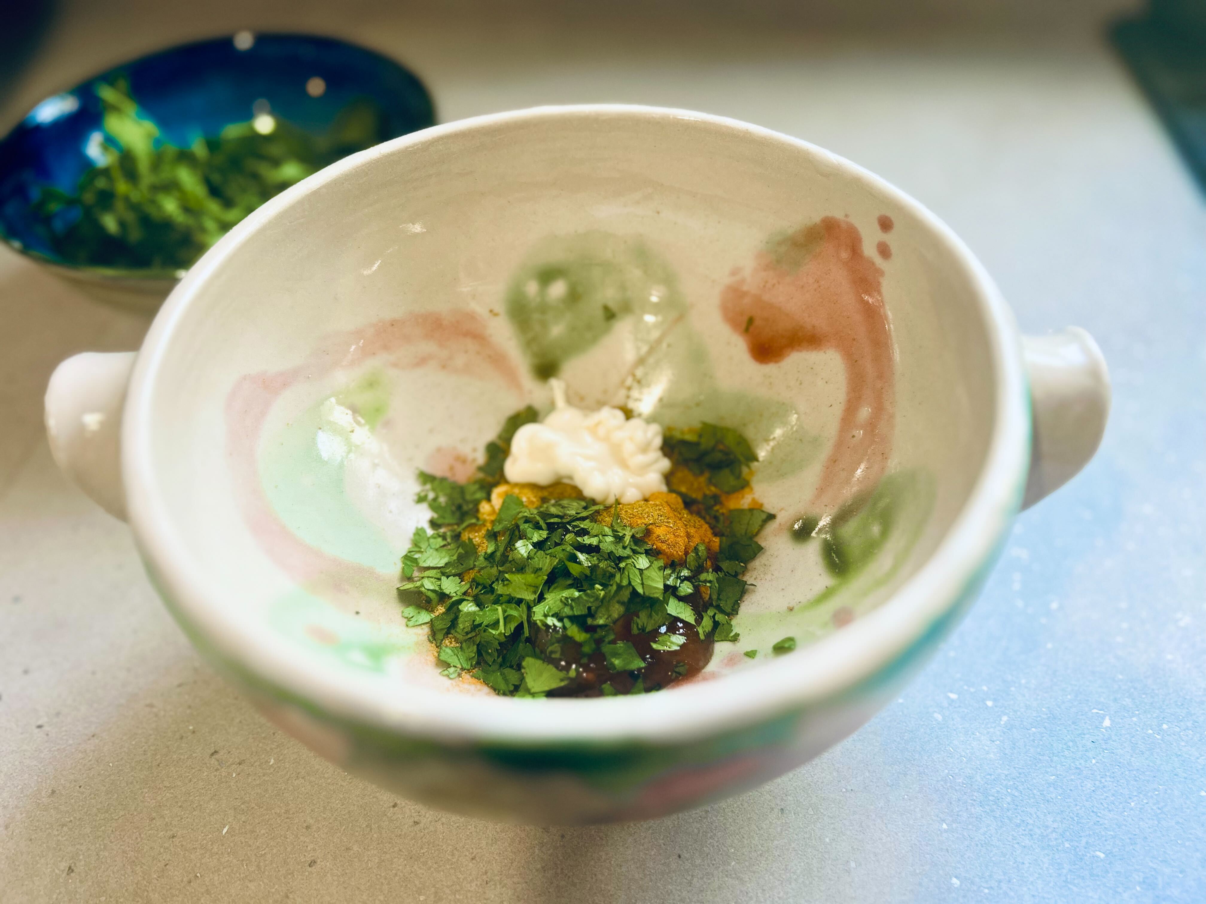 A close-up of a white, handmade ceramic bowl with pale green and pink brushstrokes. The bowl contains several ingredients for a sauce, including white mayonnaise, a small mound of dark red chutney, a pile of green chopped coriander or parsley, and a scoop of yellow curry powder. In the background, a small blue bowl filled with more green herbs sits on a marble countertop.