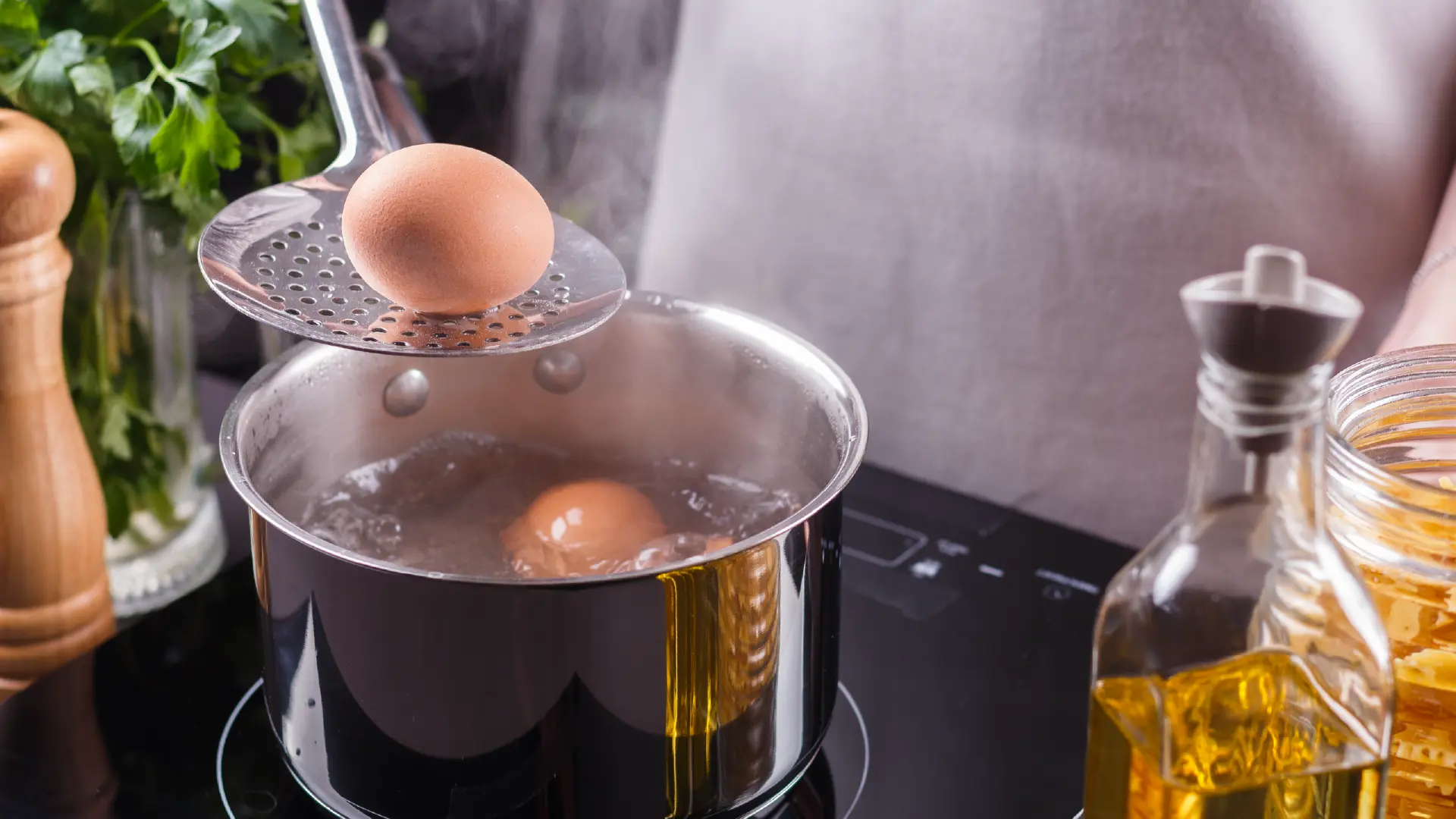 A person uses a slotted spoon to lower a brown egg into a pot of boiling water on a sleek black induction stovetop.