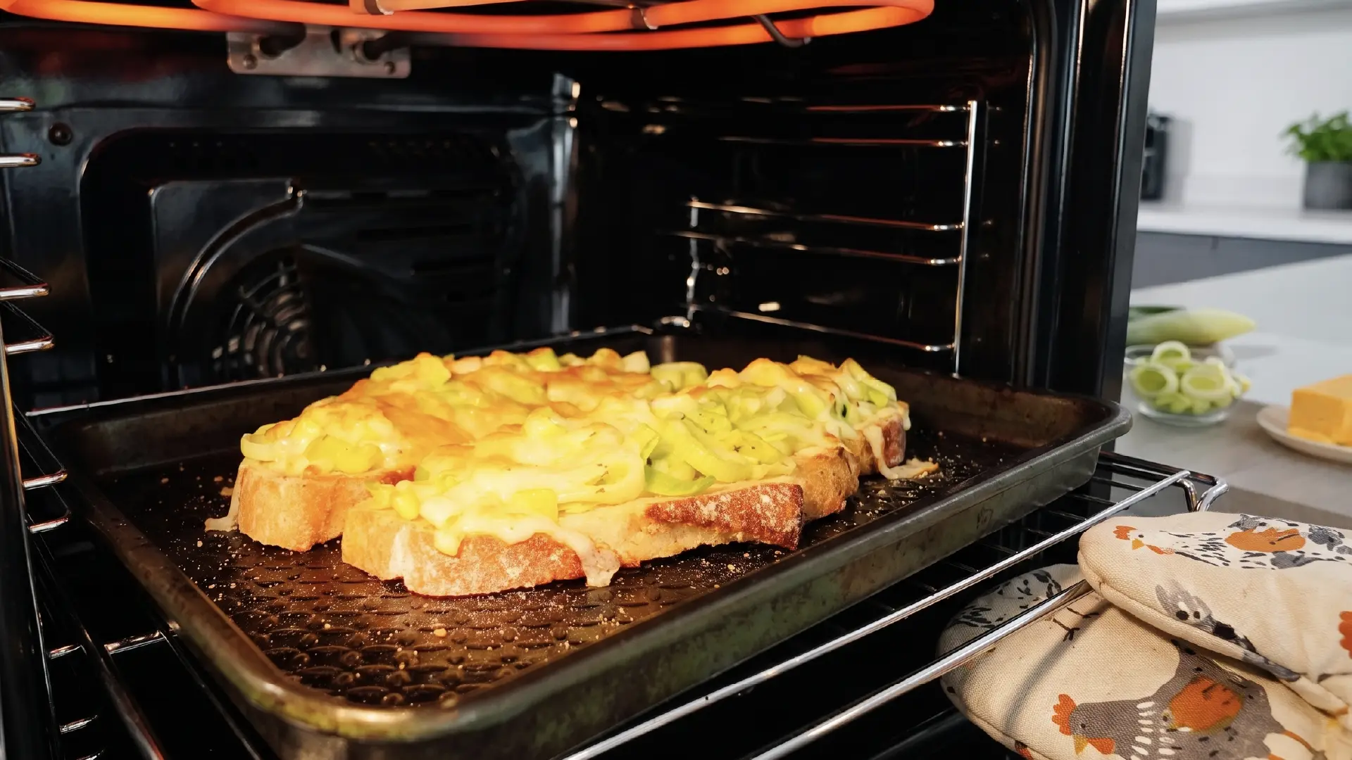 Bread topped with melted cheese and leeks bakes on a tray inside an open oven under a glowing heating element.