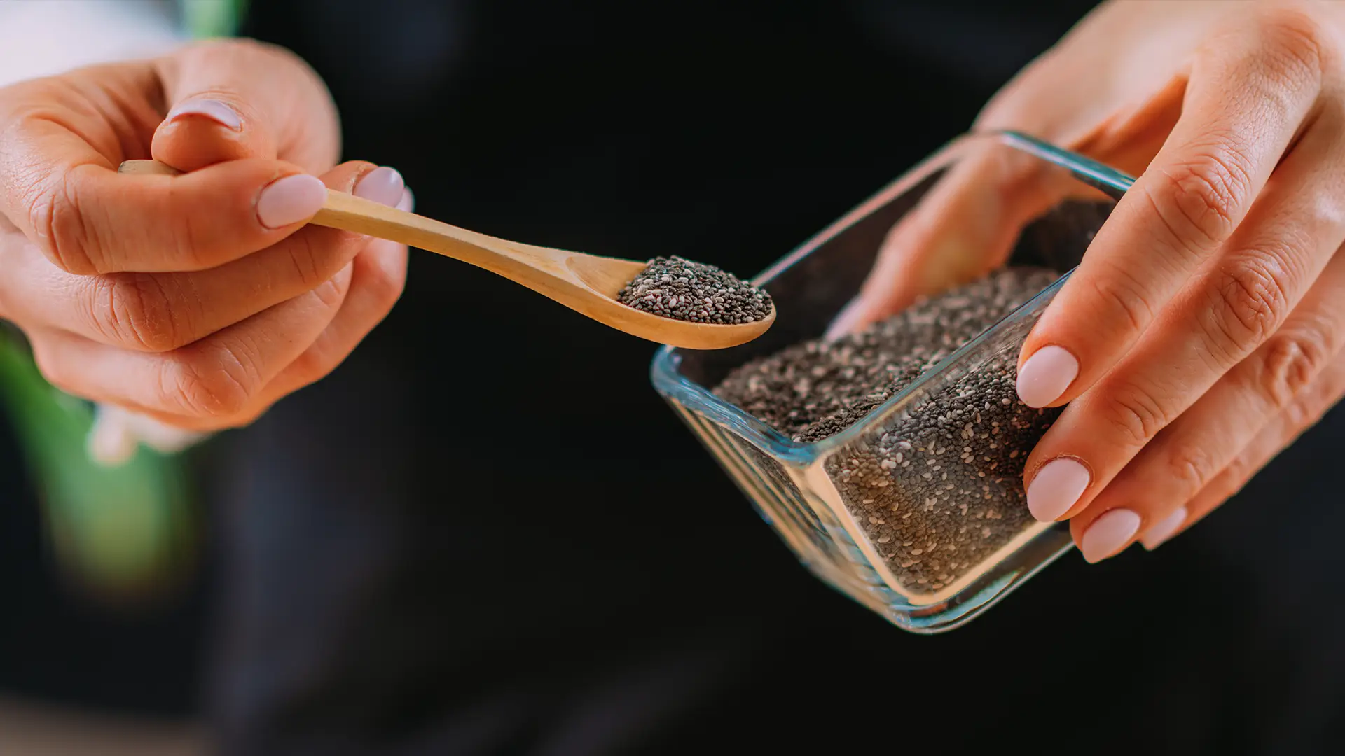 A close-up shot shows a person's hands in the foreground, with pale pink manicured nails. The left hand holds a small wooden spoon, carefully transferring black chia seeds from a clear glass rectangular container, held by the right hand, into a clear glass bowl below. The bowl already contains a mixture of chia seeds and possibly rolled oats. The person is wearing a dark apron or top, and a wooden surface is visible in the foreground.