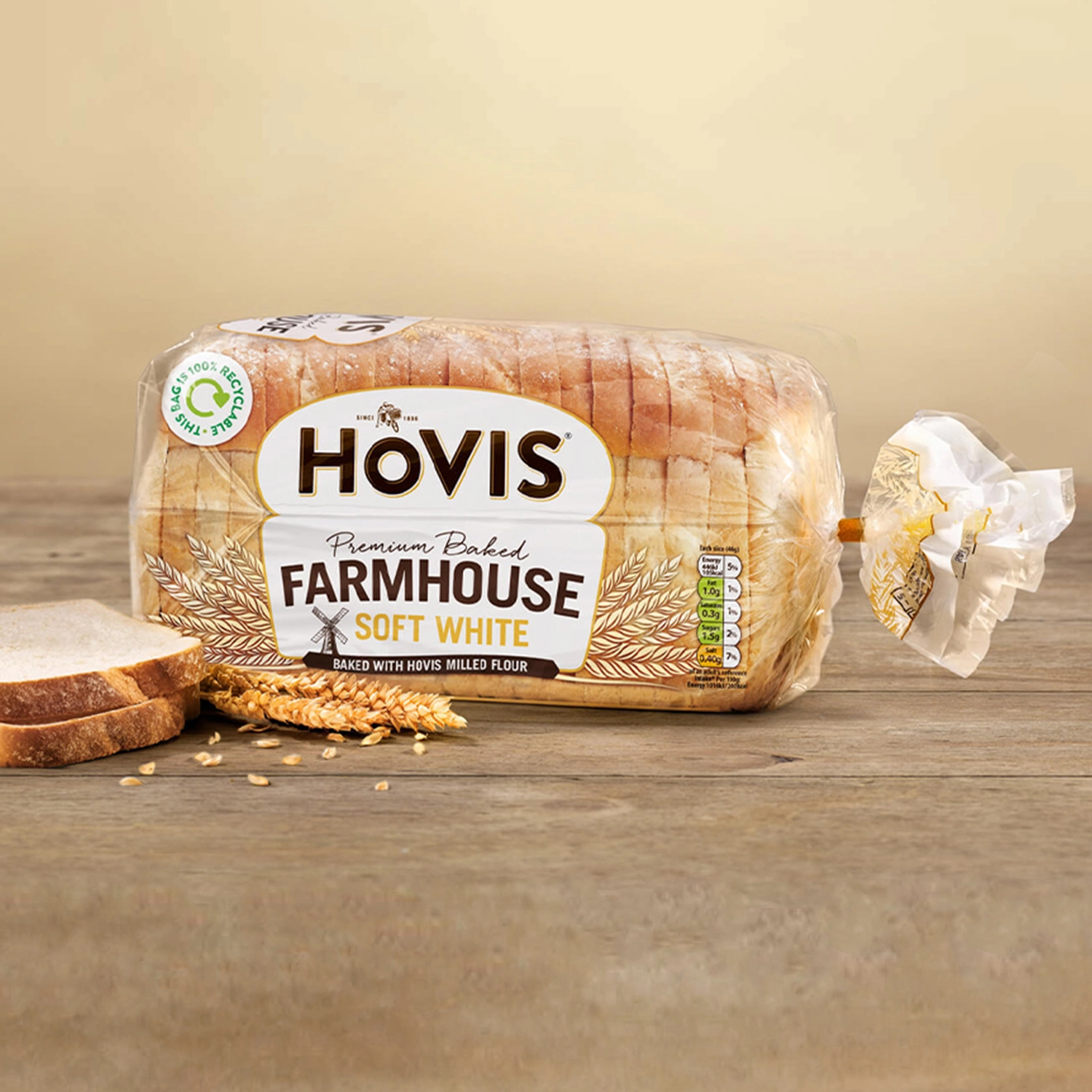 Hovis Farmhouse Soft White bread against a light beige background on a wooden table, with two stacked slices of bread on the left, alongside some wheat.