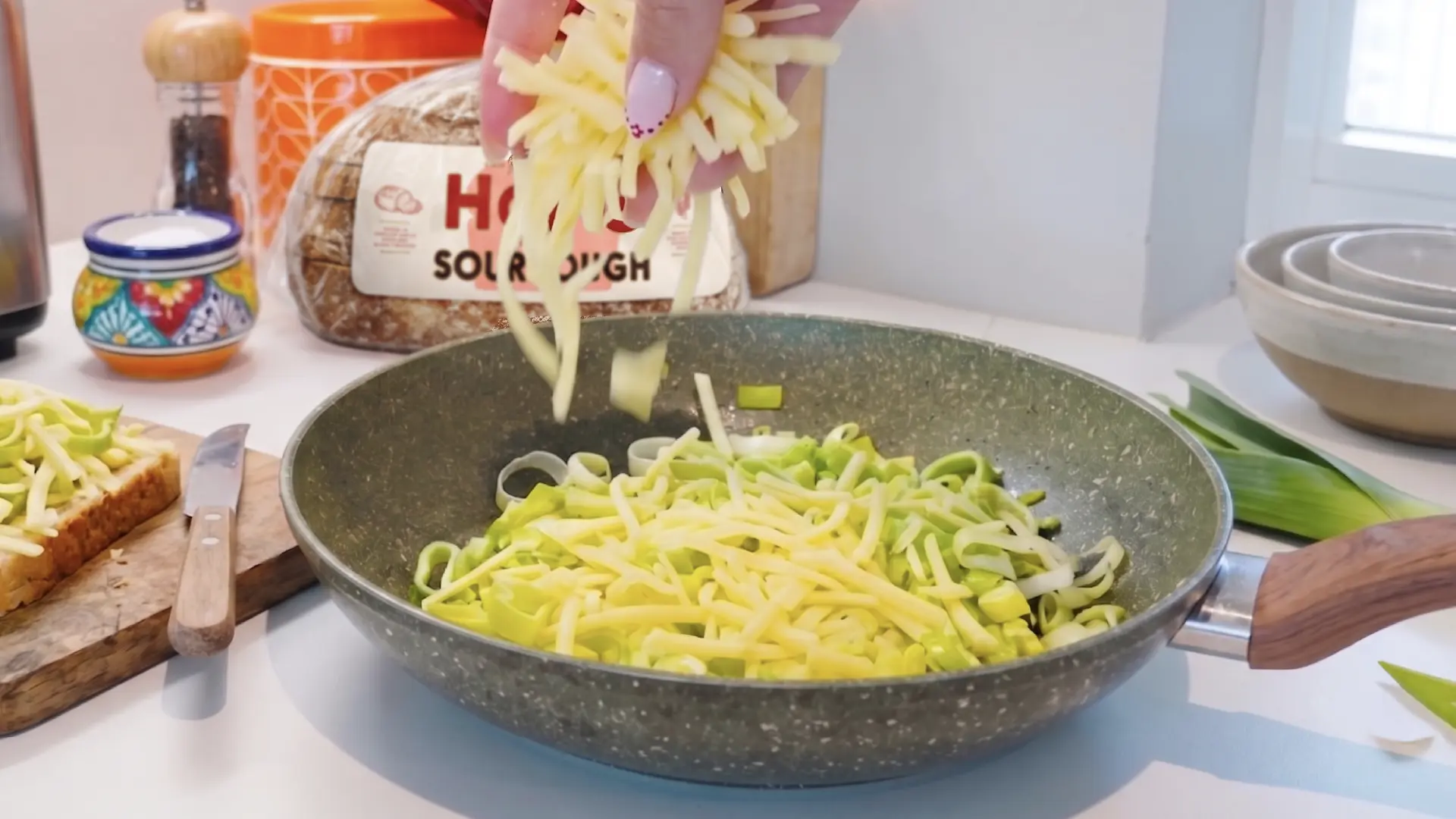 A hand sprinkles grated cheese into a non-stick frying pan filled with chopped green leeks on a kitchen counter.