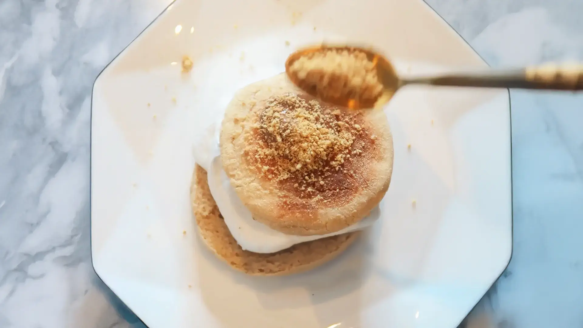 A top-down view of a spoon full of Hovis Digestive Biscuits crumbs being sprinkled on top of the English muffin Smores, on an octagonal plate. 