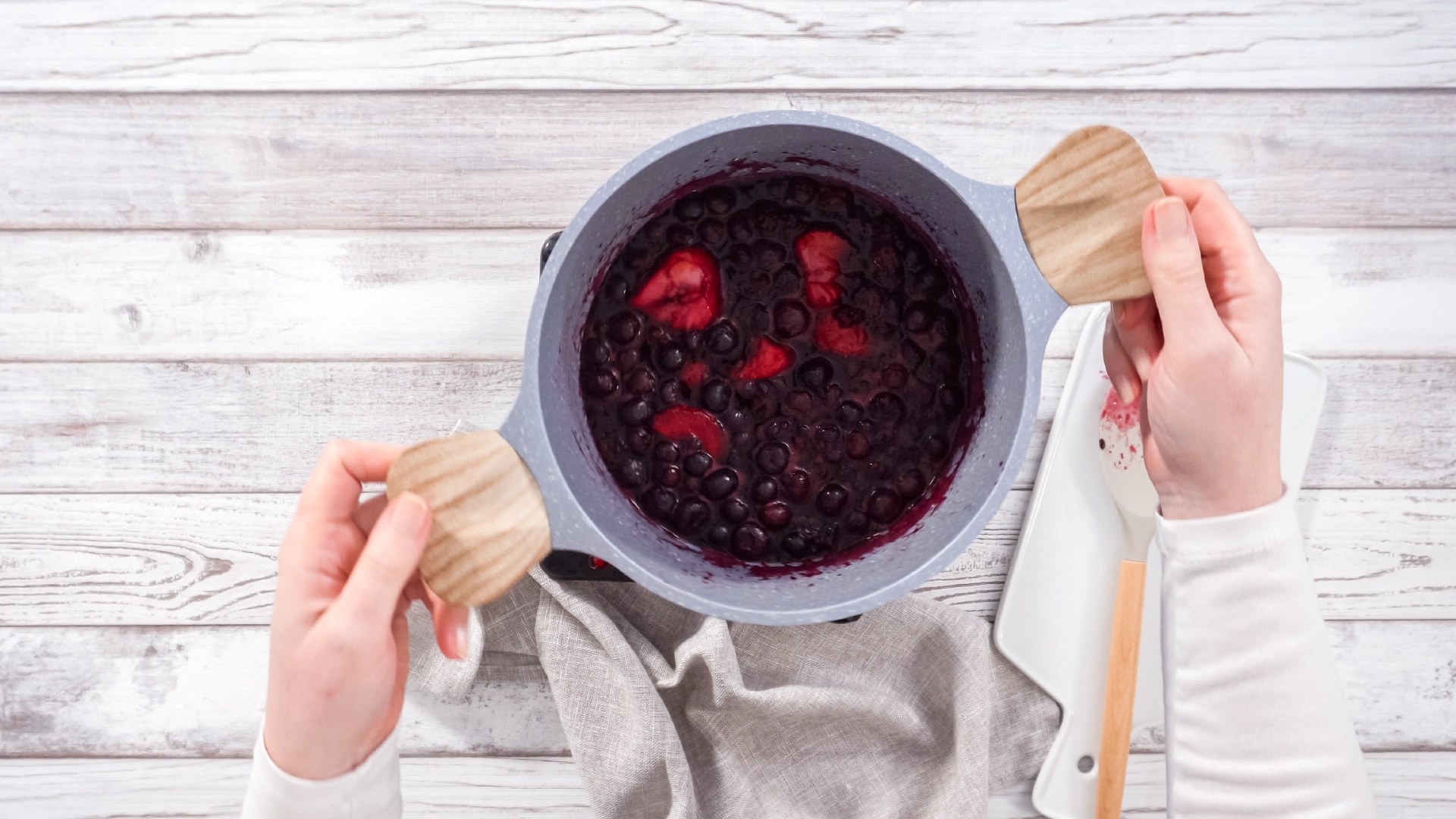A top-down view of a person using both hands to hold a medium pot filled with red berries and purple juice on a wooden table.