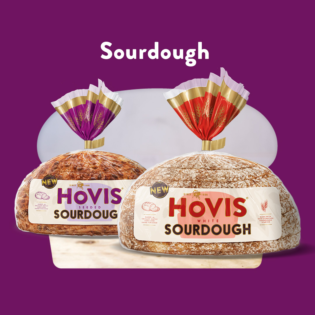Two loaves of Hovis sourdough bread are displayed against a purple background. One loaf is labelled “Hovis Seeded Sourdough” in purple packaging, and the other is labelled “Hovis White Sourdough” in red packaging. Both loaves are round, crusty, and wrapped in clear plastic with gold and coloured tops. The word “Sourdough” appears above them in white text.