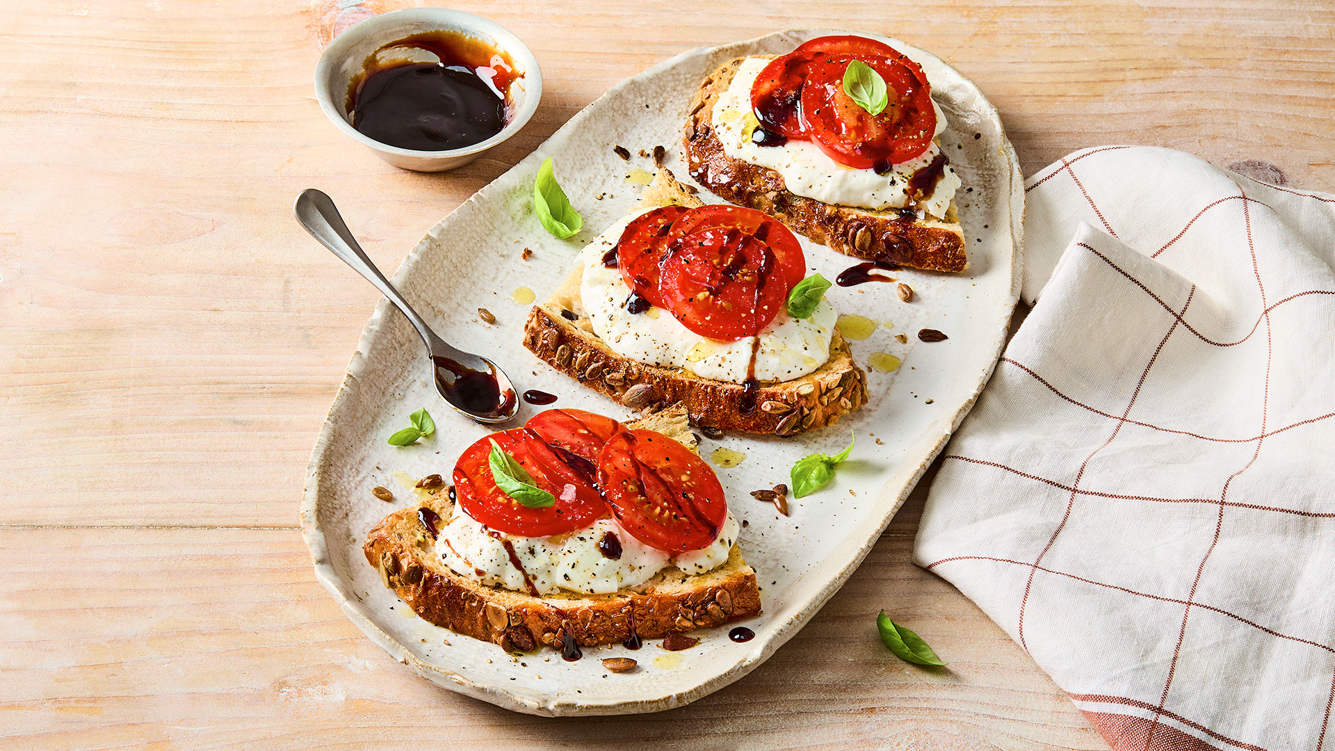 3 half slices of seeded sourdough on a plate topped with burrata cheese and tomato, drizzled with balsamic glaze and sprinkled with basil leaves. There is a spoon on the plate next to a bowl of extra balsamic glaze