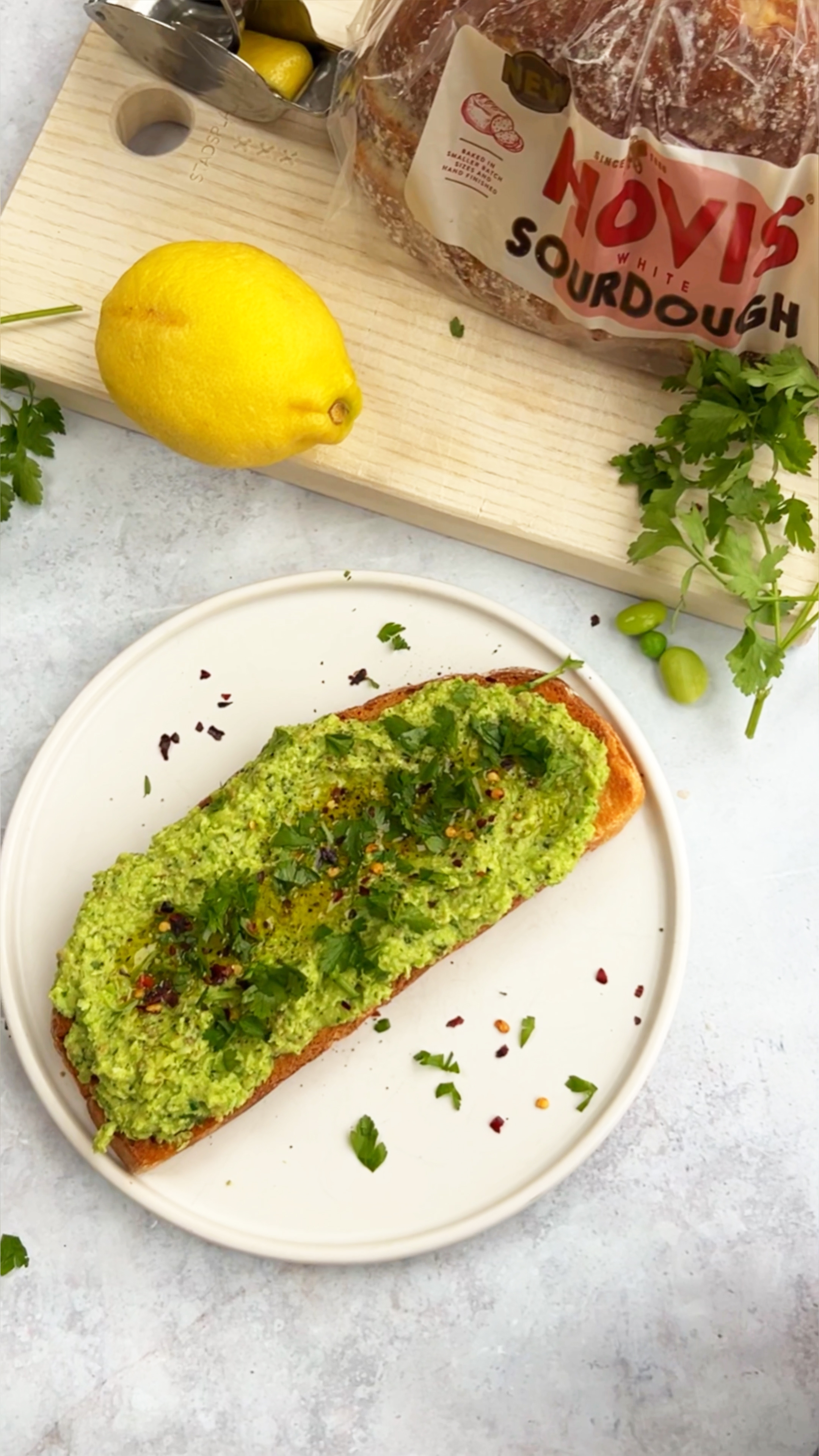 A slice of toasted sourdough is topped with a vibrant green pea and herb spread, garnished with fresh parsley, chilli flakes, and a drizzle of olive oil. The toast sits on a simple white plate, surrounded by scattered herbs. In the background, a loaf of Hovis White Sourdough rests on a wooden board alongside a lemon and a metal lemon squeezer, adding a bright, fresh feel to the scene.
