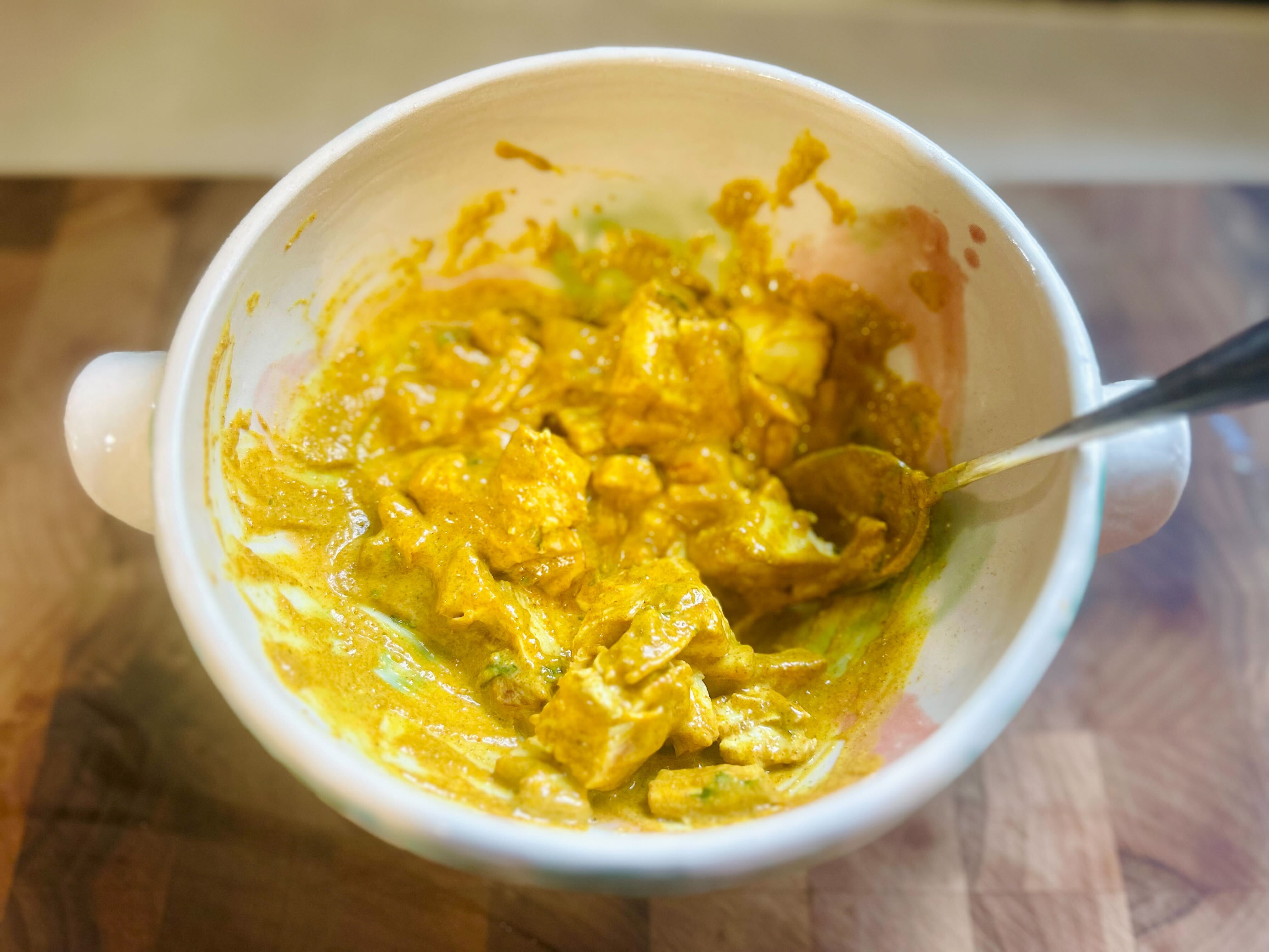 A close-up, top-down shot of a ceramic bowl on a wooden chopping board. A metal spoon is stirring together large chunks of cooked chicken with a thick, yellow Coronation Chicken sauce. The sauce coats all the pieces.