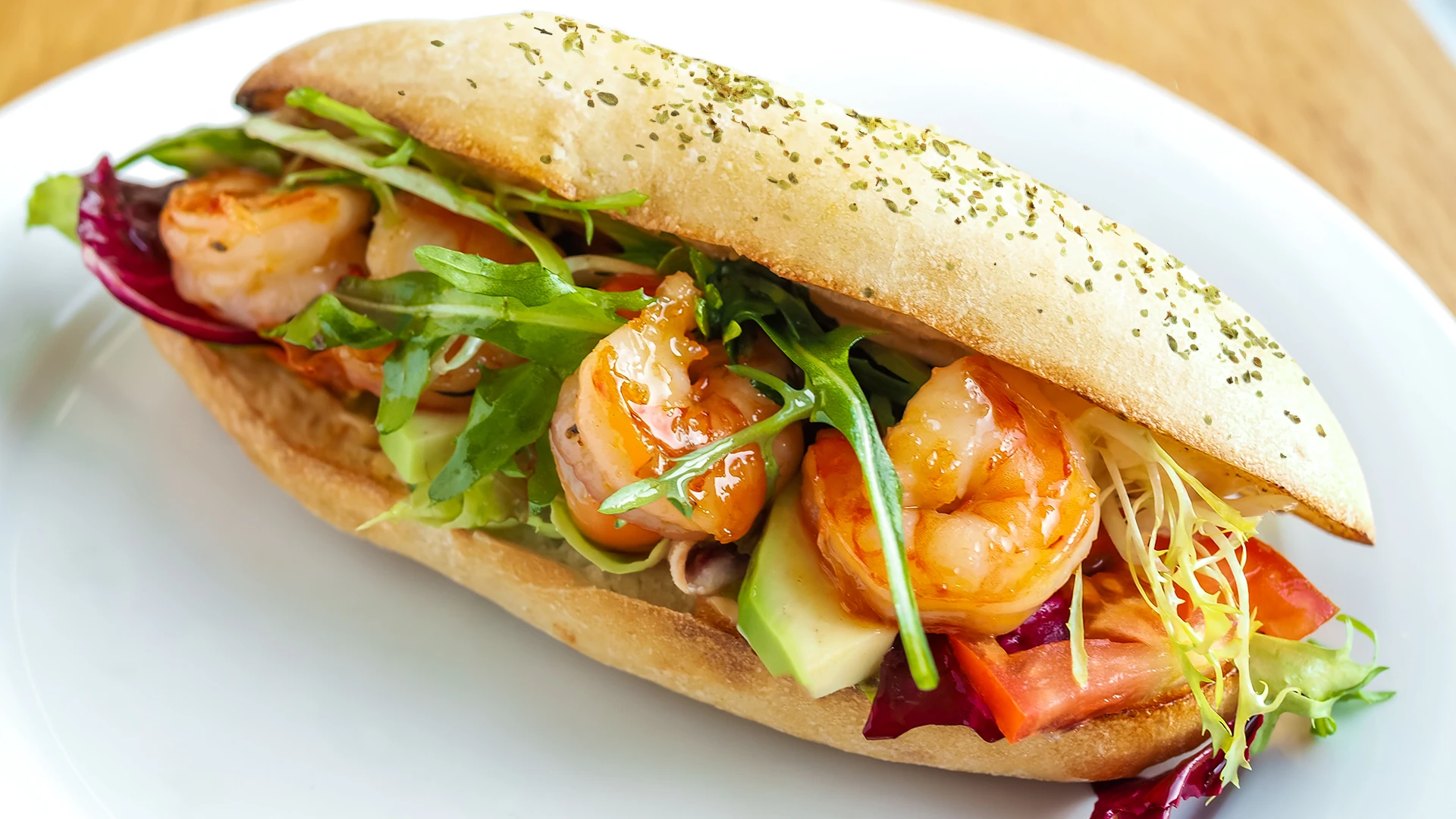 A full Spicy Prawn Sandwich sits on a white plate on a wooden kitchen counter filled with avocado, lettuce and prawns