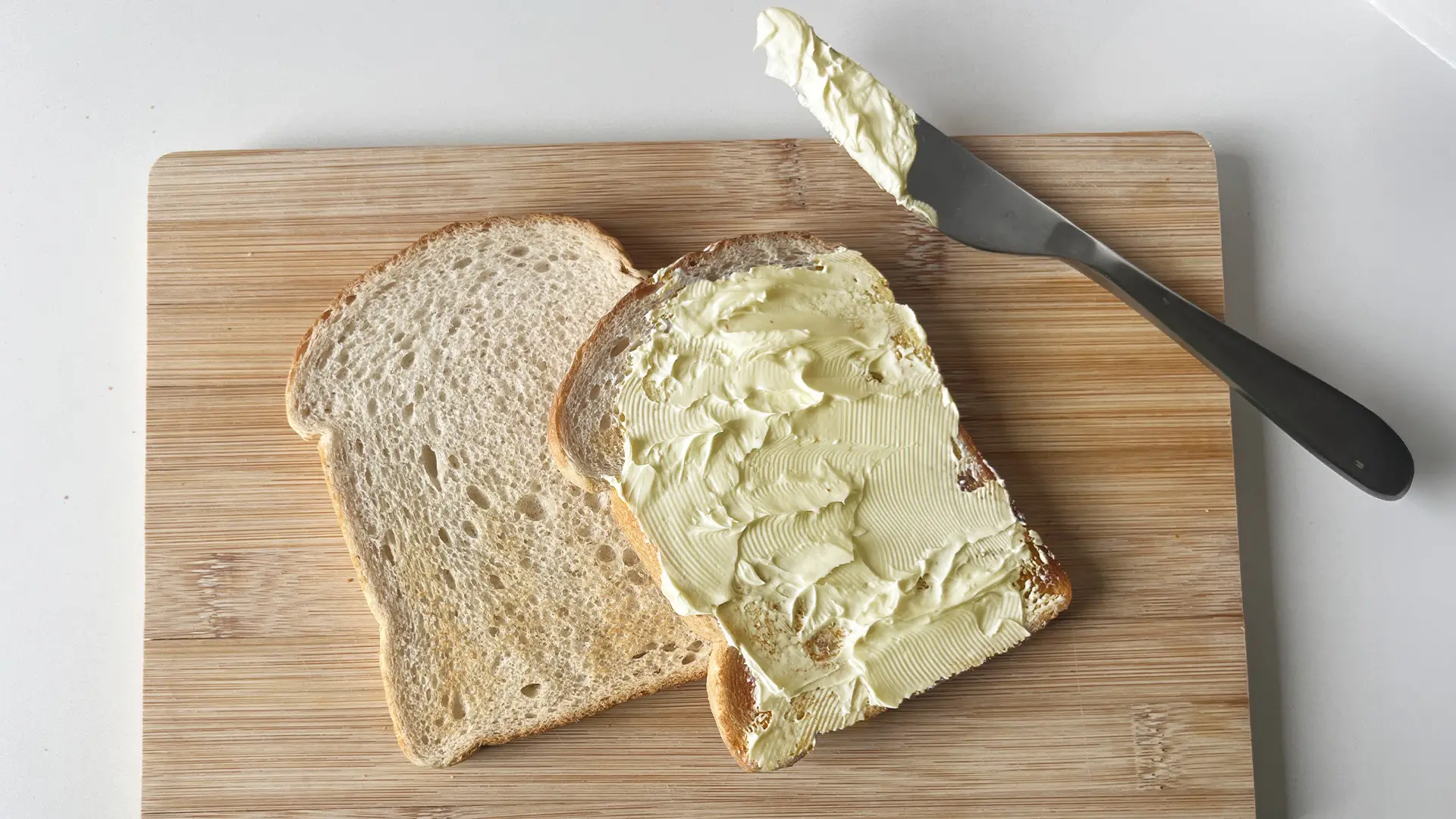 A top-down view shows two slices of lightly toasted bread on a light wooden cutting board. The slice on the right is generously spread with butter or a light-coloured spread, showing visible knife marks. The slice on the left is plain. A silver butter knife with a dark handle rests on the board above the spread toast, with a dollop of spread still on its blade. The cutting board is positioned on a plain white surface.
