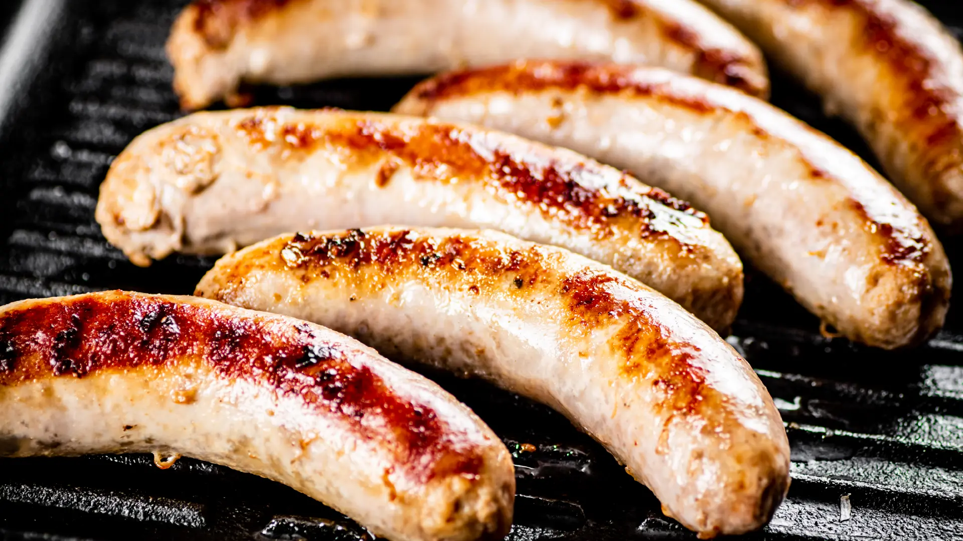 Six juicy bratwurst sausages grilling on a black ribbed stove top, showing deep golden-brown char marks.