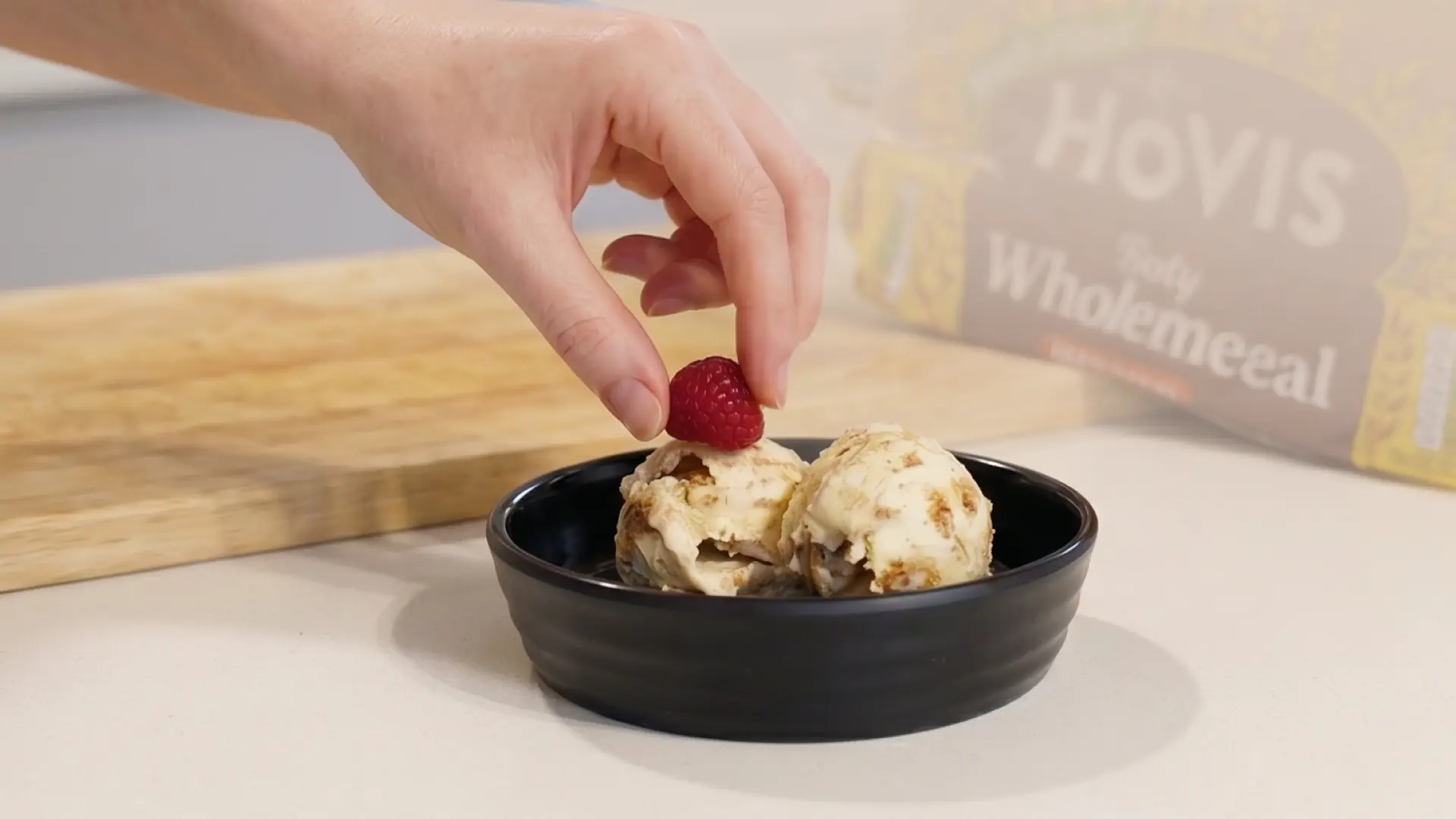 A hand places a raspberry on two scoops of ice cream in a black bowl, with a Hovis bread loaf blurred in the background.