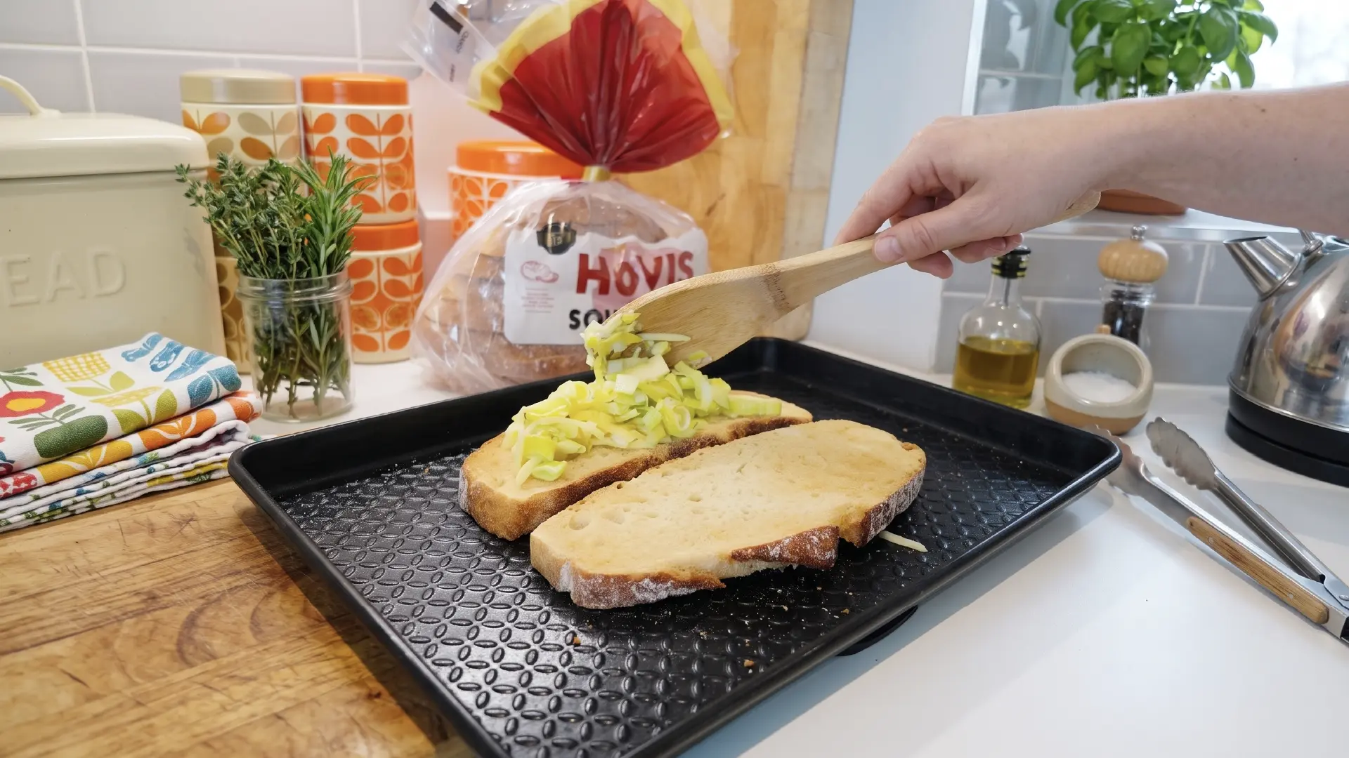 A hand uses a wooden spatula to spoon a leek and cheese mixture onto thick bread slices on a black baking sheet.