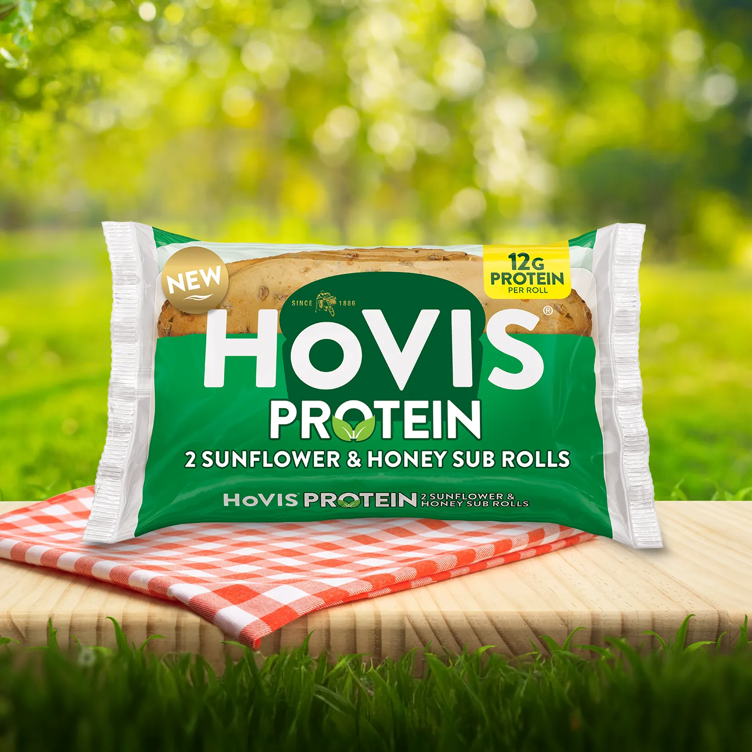Hovis Protein Sunflower Honey Sub Rolls packaging on a picnic table with a green outdoor background. The pack shows two soft sub rolls with delicious sunflower seed on top “Deliciously soft and flavourful with a hint of honey.” A red gingham cloth adds a picnic feel to the setting.