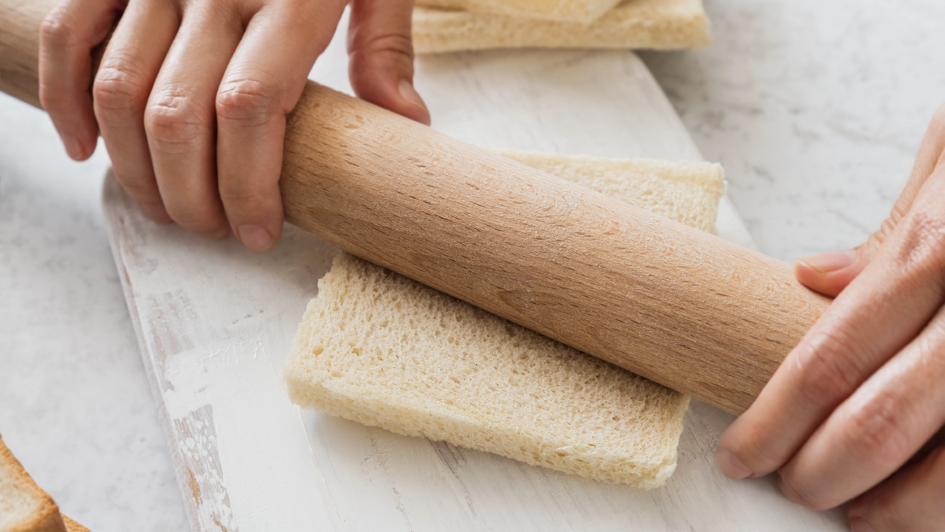 Someone uses a wooden rolling pin to flatten crustless slices of Hovis Soft White Thick Sliced on a white chopping board.