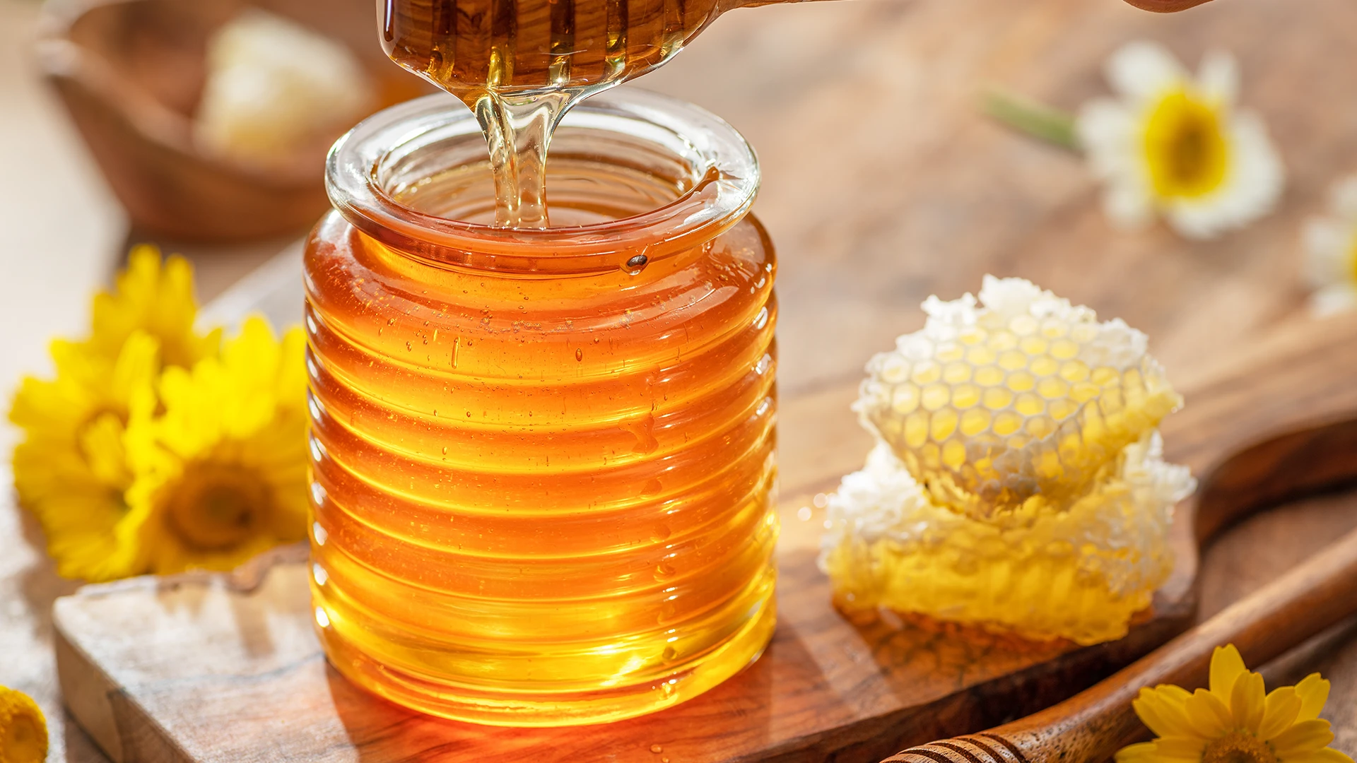 A hand lifting a honey drizzler filled with honey from a glass honey pot on a wooden chopping board, with pieces of honeycomb on the side.