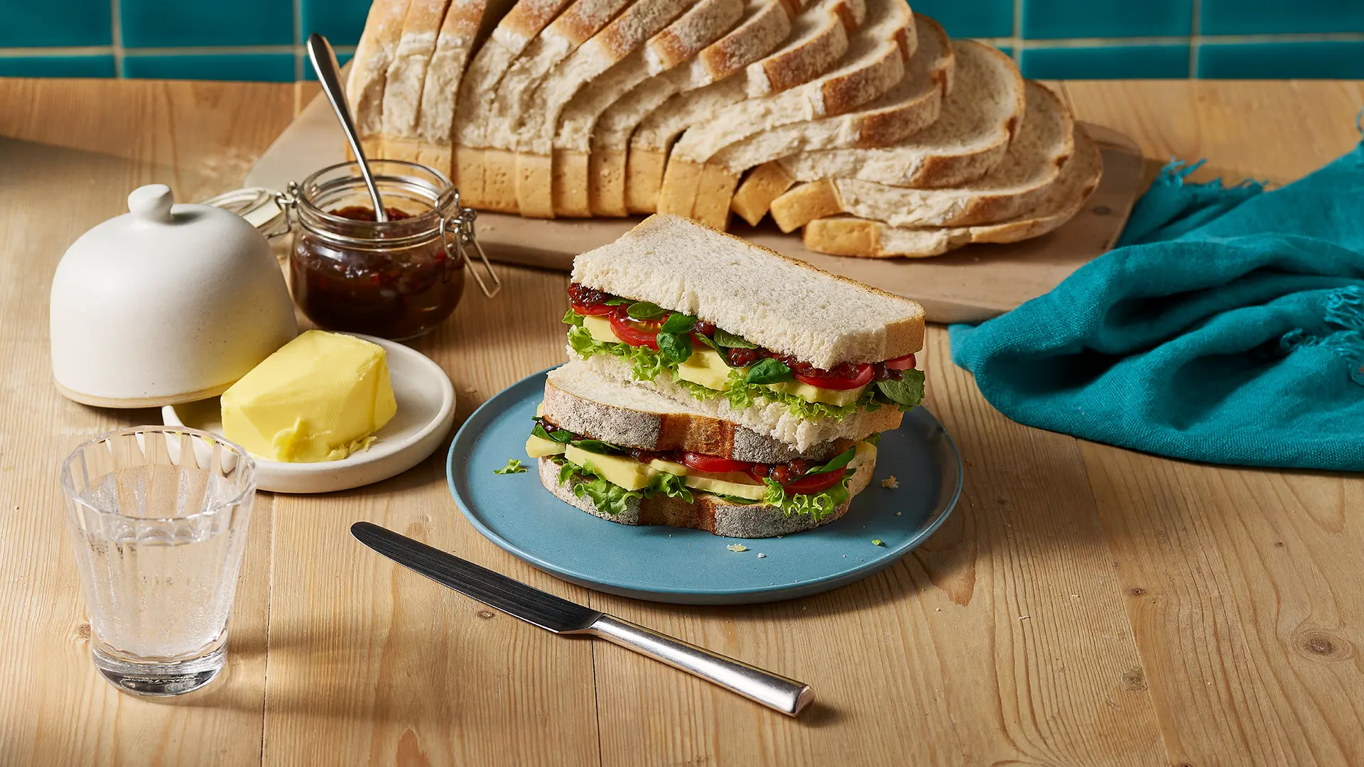 Two halves of a Hovis Farmhouse Batch sandwich filled with cheese, onion chutney, lettuce leaves, sliced tomatoes, and basil leaves. In the background, the rest of the loaf, a jar of onion chutney, and some butter.