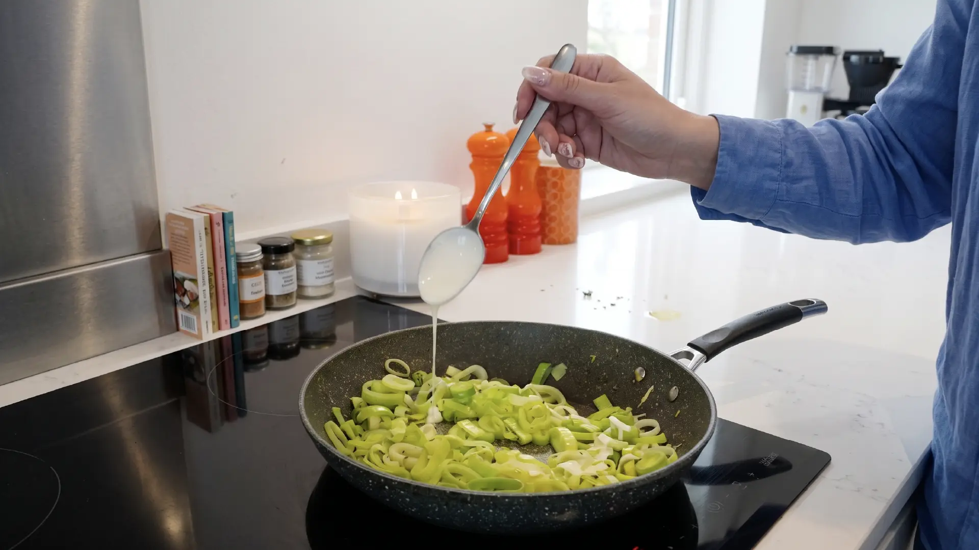 A person in a blue shirt uses a metal spoon to drizzle a white sauce over chopped leeks cooking in a frying pan.