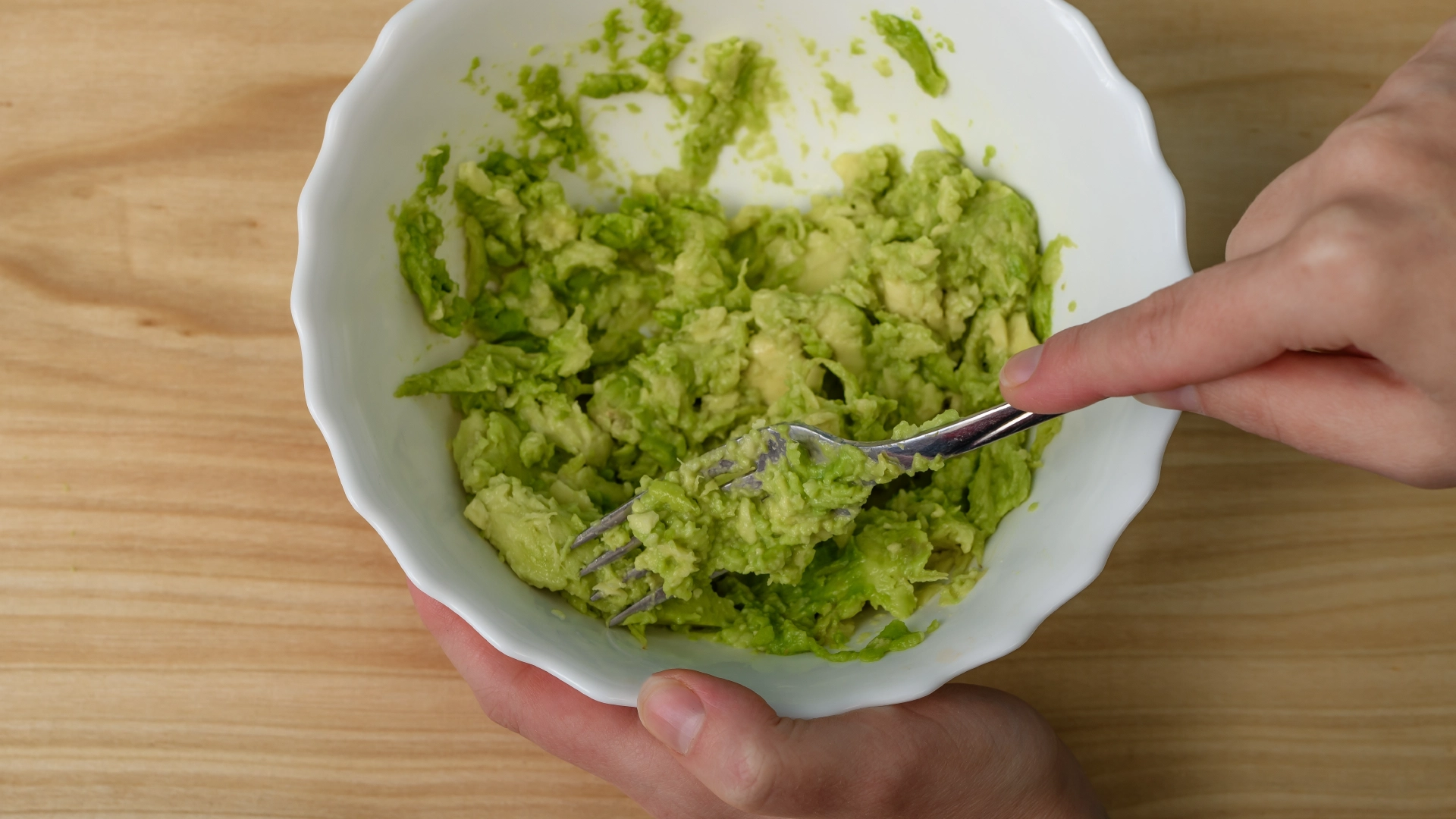 Hand holding a small porcelain bowl of avocado being mashed with a metal fork on a wooden table.