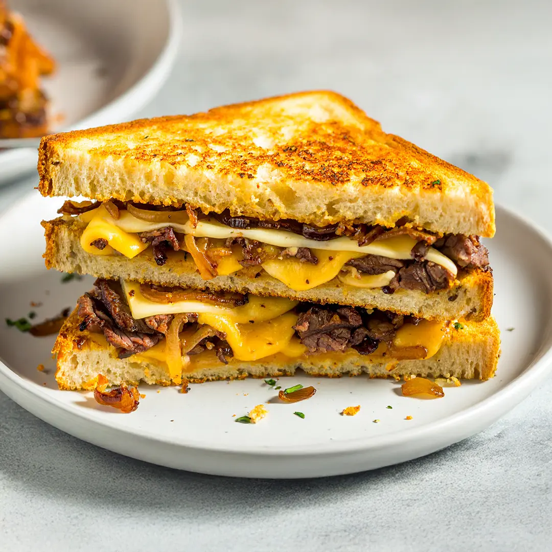 A delicious Beef & Caramelised Onion Grilled Cheese sandwich cut in half on a white plate, on a marbled table. In the background, another sandwich on a plate. 