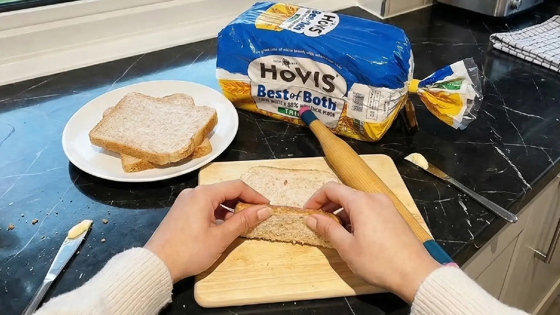 Hands rolling a slice of Hovis bread on a wooden board next to a rolling pin, butter knives, and a loaf of bread.