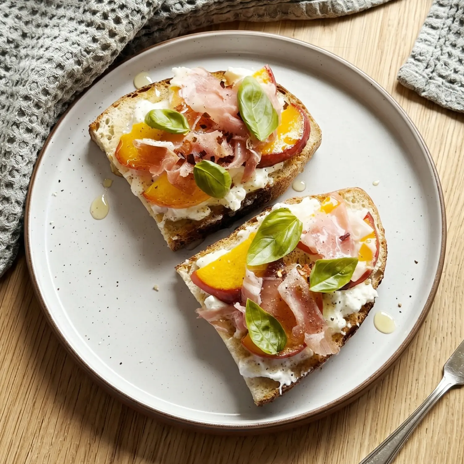 Two slices of artisan toast topped with white cheese, yellow peaches, prosciutto and fresh basil leaves on a plate.