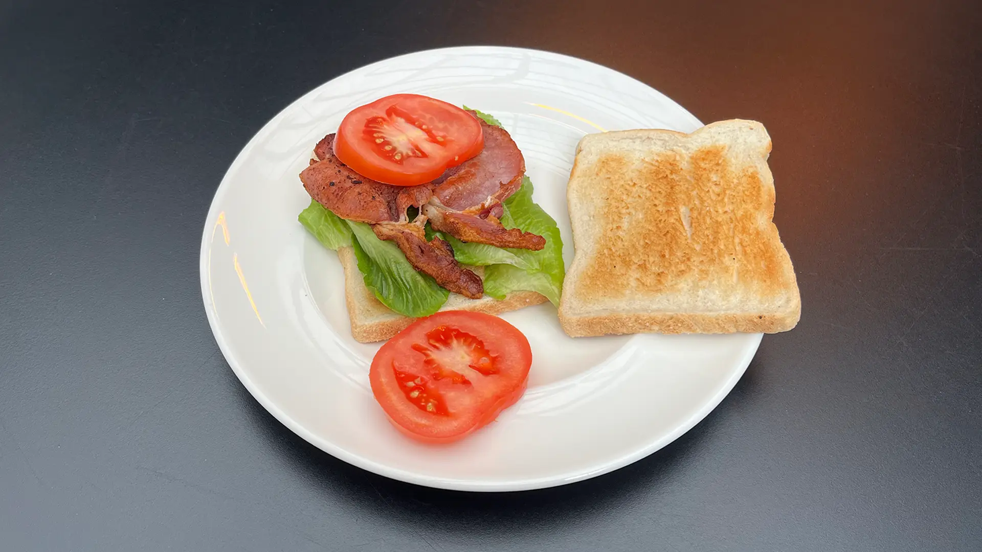 A hand holds a slice of bacon while the other slices it in half with a knife on a wooden chopping board. In the background, there are stacked slices of bread, lettuce leaves, and tomato slices.
